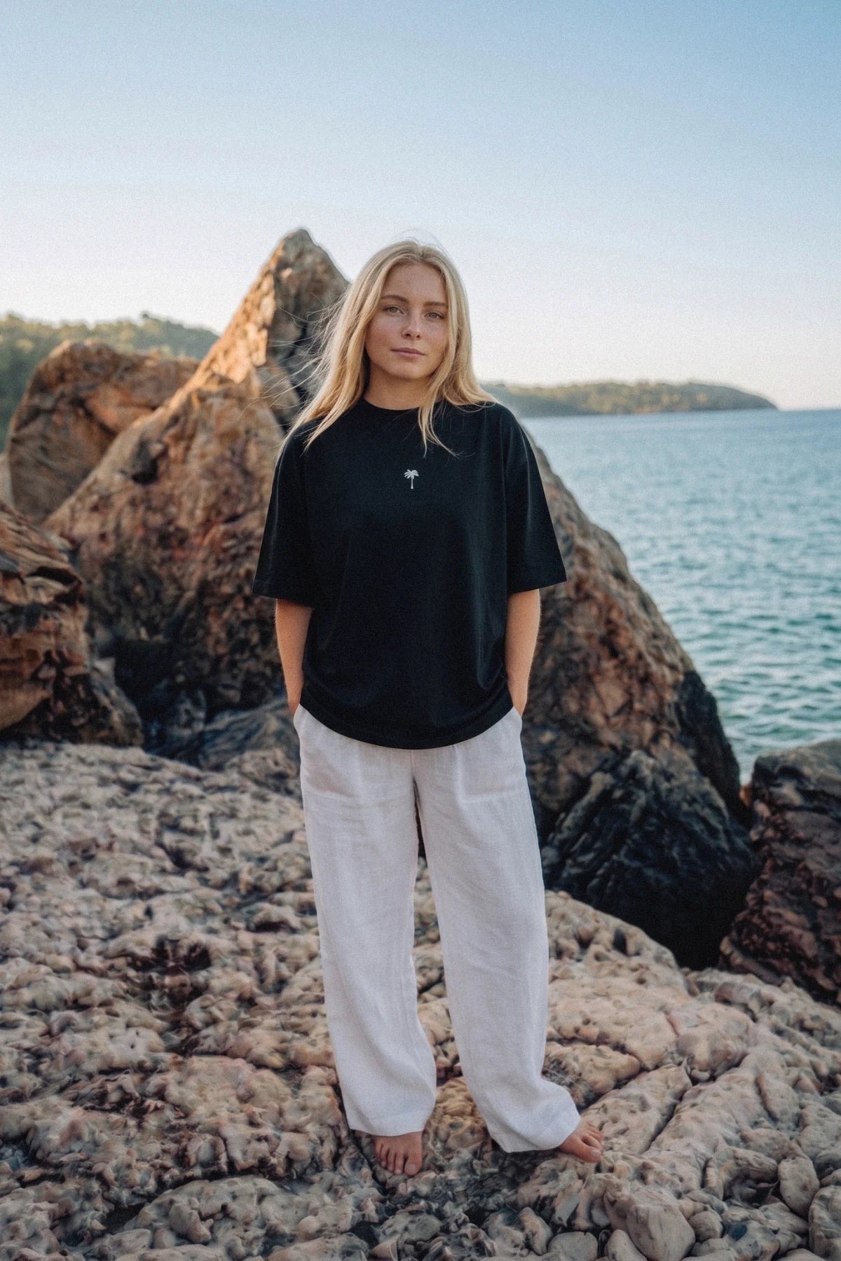 Schwarzes Palm Tree Boxy Oversized T-Shirt für Damen mit gesticktem Palmenlogo auf der Brust, kombiniert mit weiter Leinenhose an einer felsigen Küste.