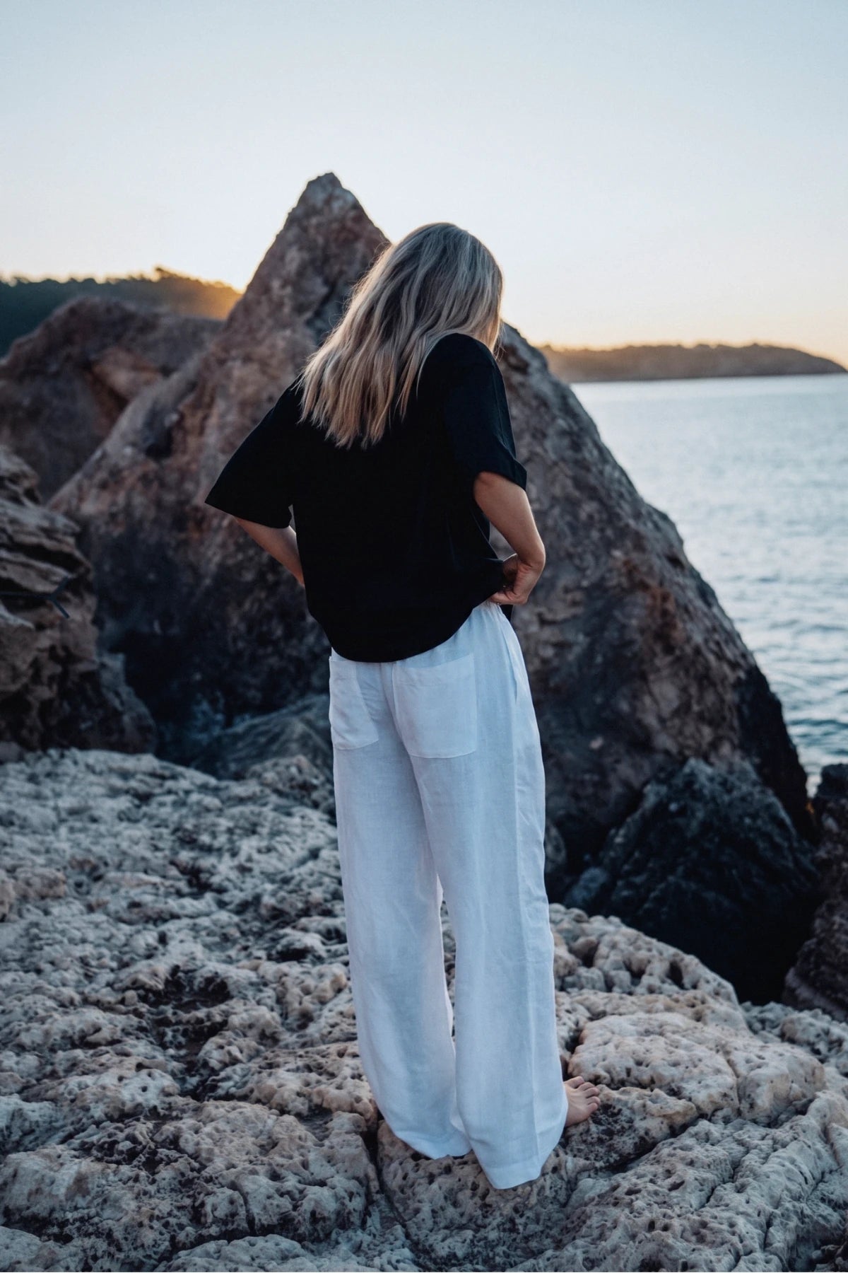 Rückansicht des Palm Tree Boxy Oversized T-Shirt Black an einer felsigen Küstenlandschaft bei Sonnenuntergang mit Blick auf das Meer.