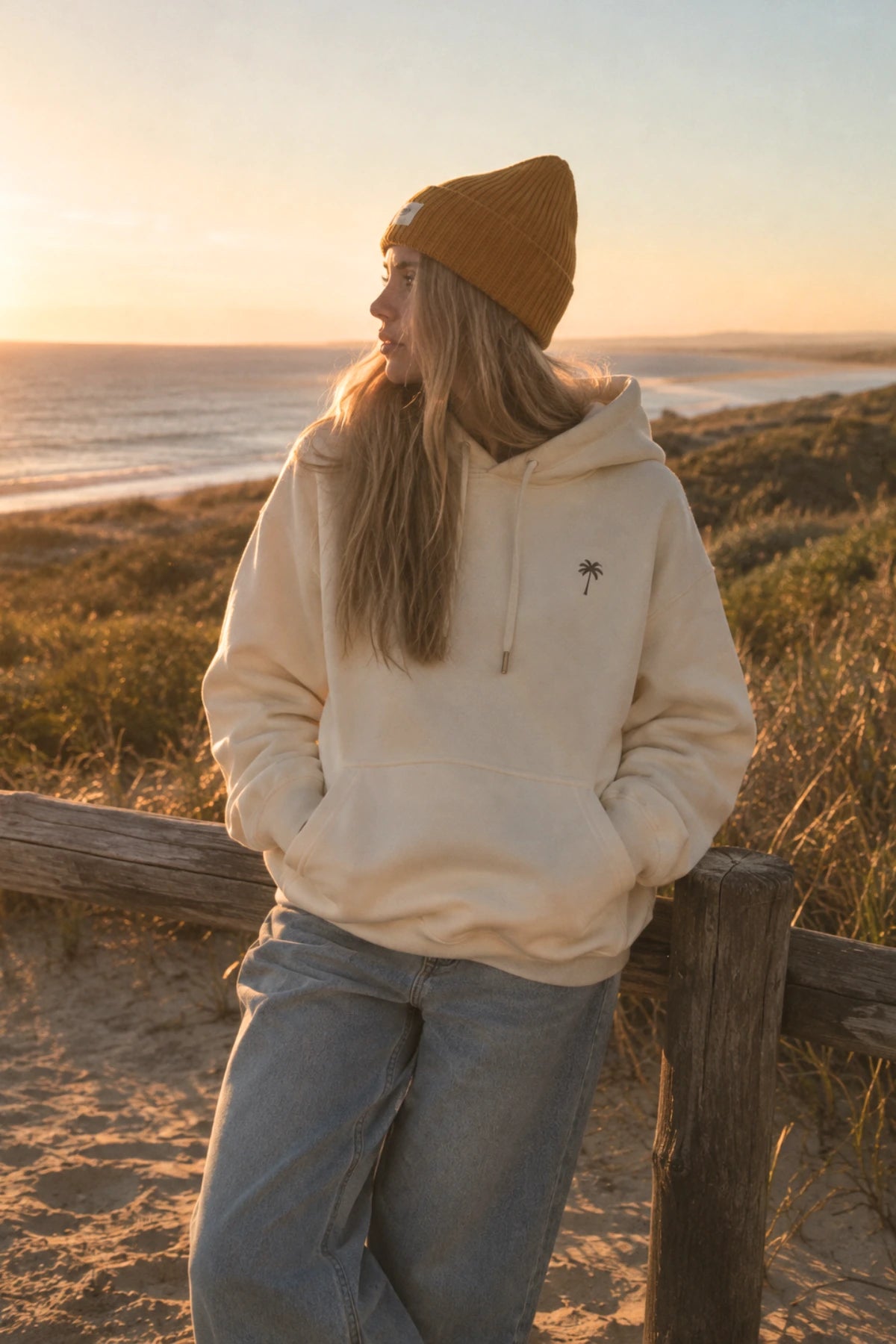 Frau am Strand mit cremefarbenem Kapuzenpullover Palm Tree Hoodie in Natural Raw von The Sunkissed Rebels, Kängurutasche und gelber Beanie.