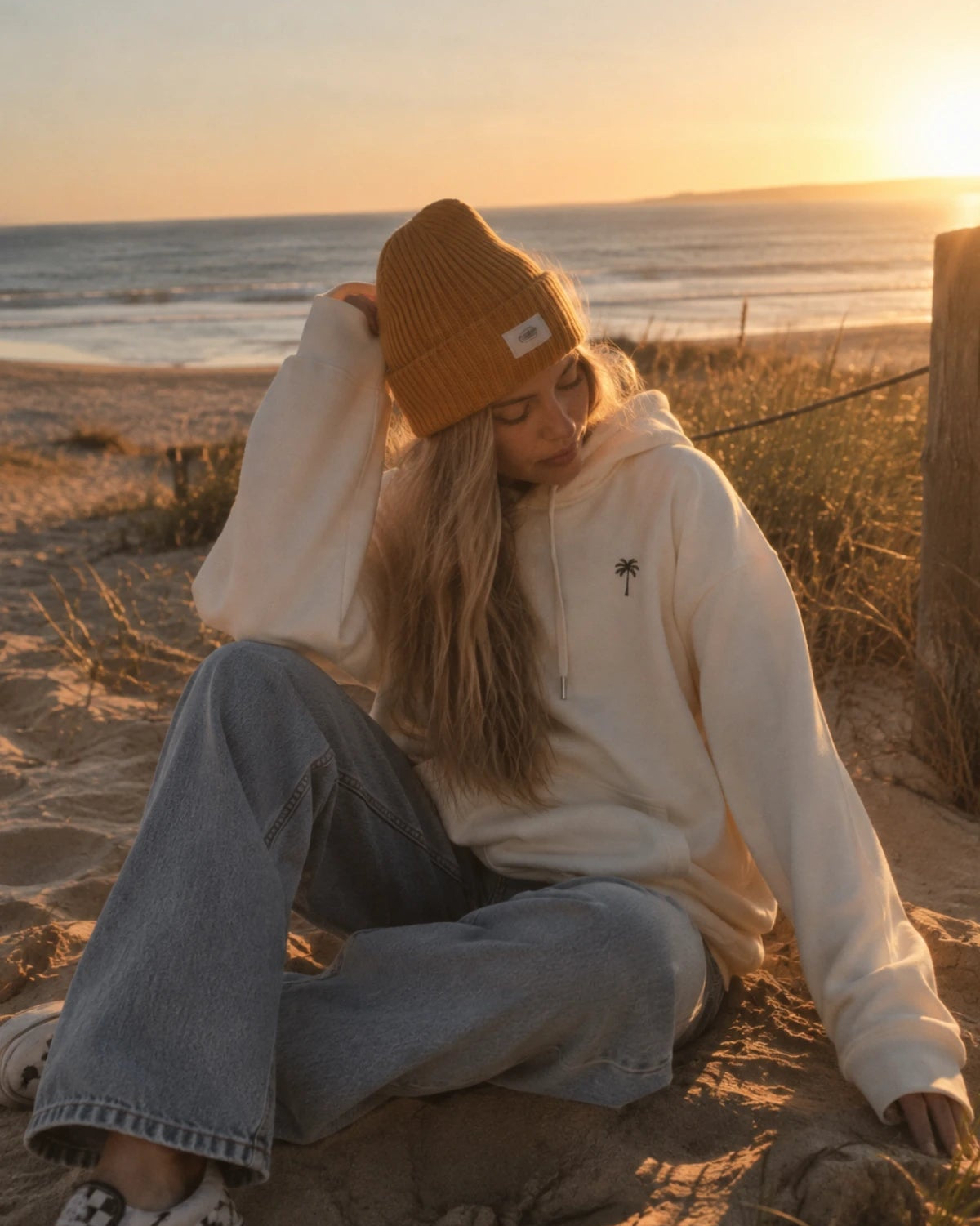 Entspannter Strandlook mit beigem Oversized Hoodie und dezenter Palmen-Stickerei von The Sunkissed Rebels im warmen Sonnenuntergang.