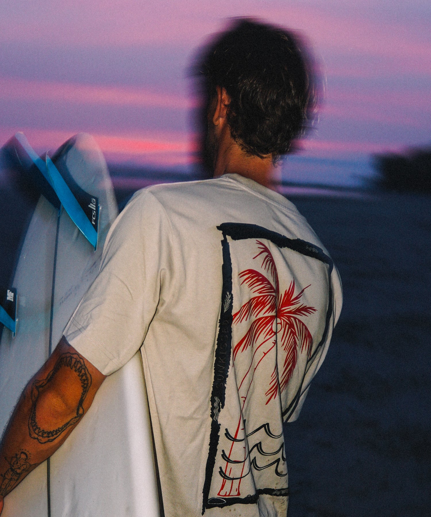 Rückansicht eines Surfers mit weißem Oversized T-Shirt und großem Palmen-Rückenprint von The Sunkissed Rebels bei Sonnenuntergang.