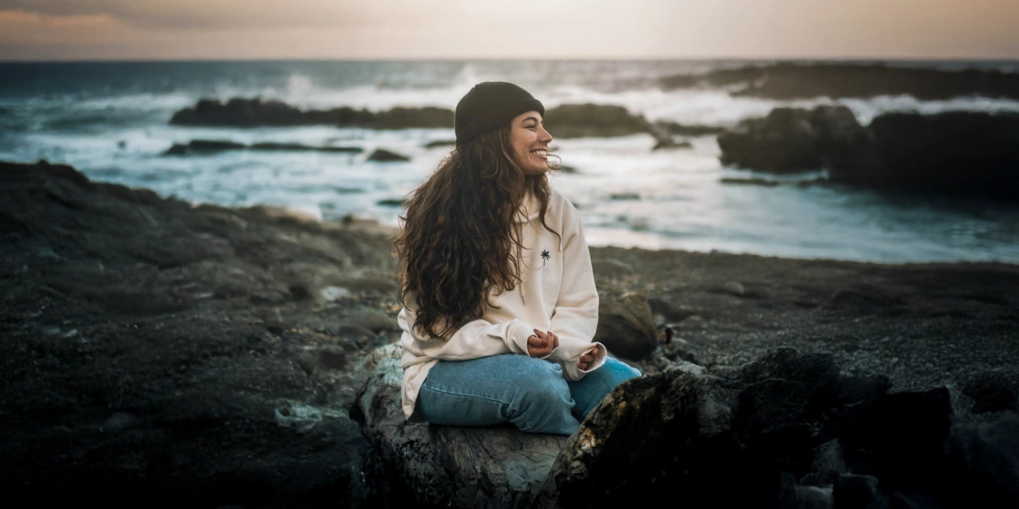 Frau mit beigem Hoodie und Palmen Stick sitzt am Strand im Winter