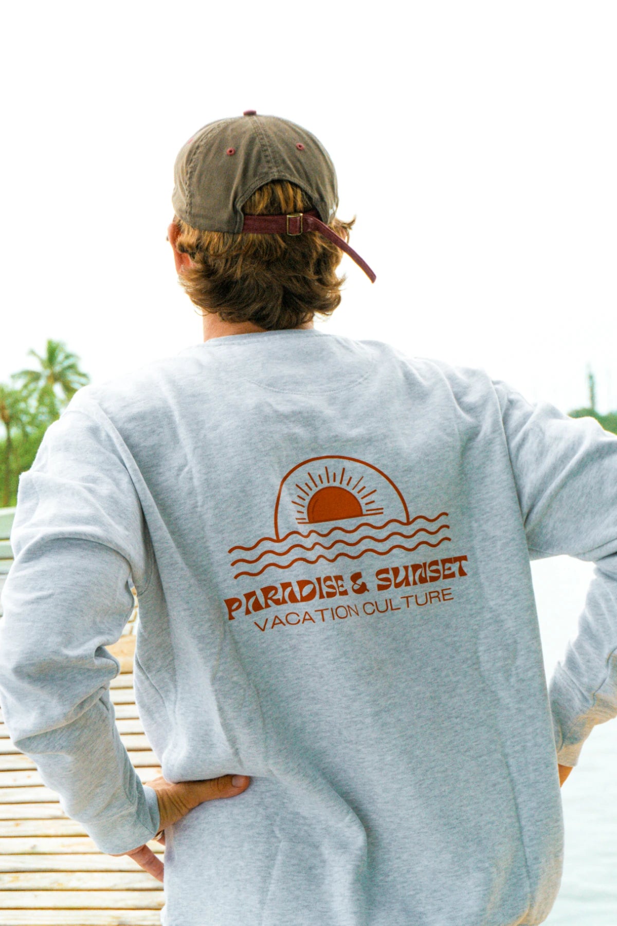 Rückansicht des Paradise & Sunset Sweaters in Cool Grey mit großem Paradise & Sunset Rückenprint auf einem Steg am tropischen Wasser.