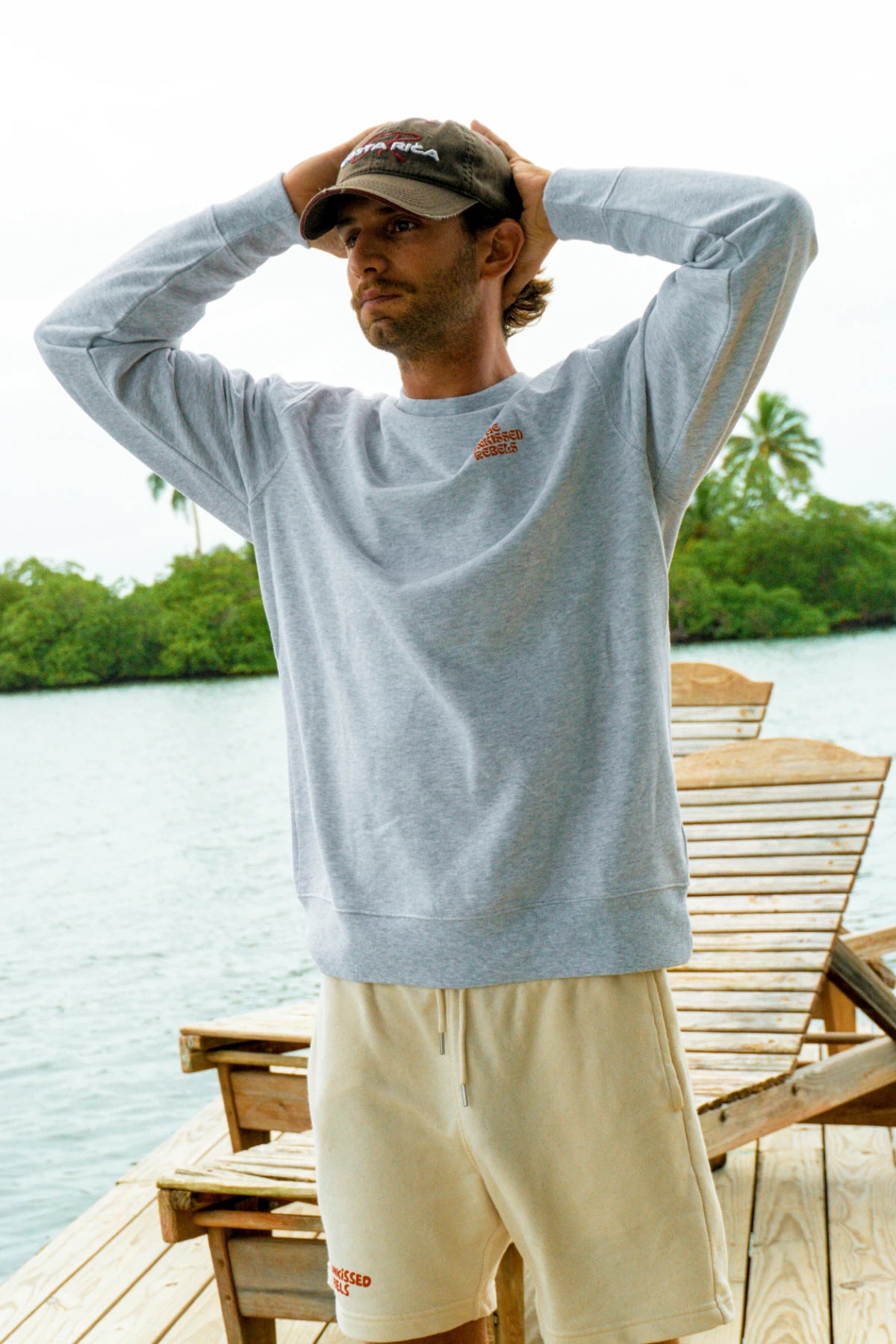 Mann trägt den Paradise & Sunset Sweater in Cool Grey auf einem Holzsteg am tropischen Wasser mit entspannter Vacation-Vibes-Atmosphäre.