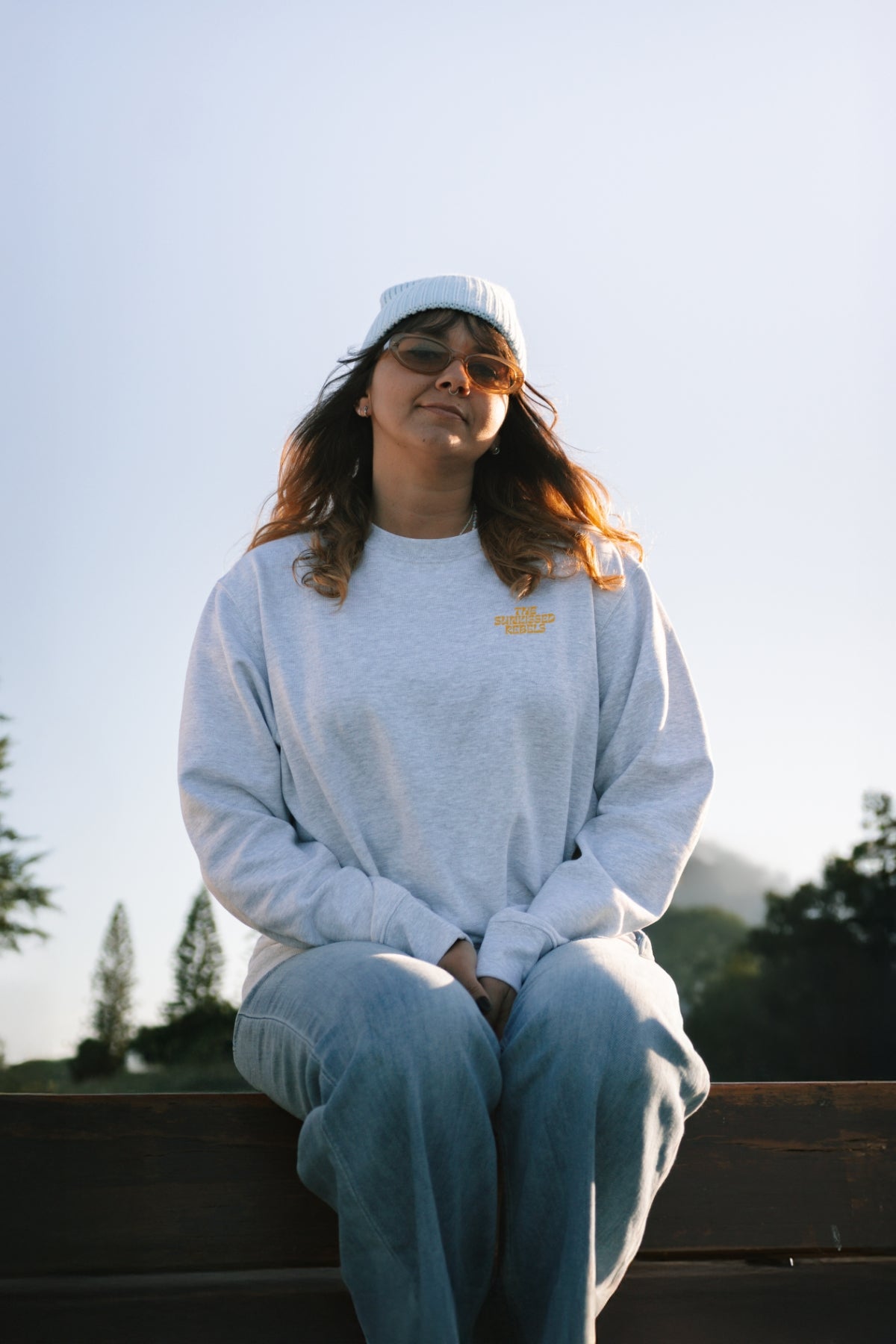 Frau sitzt auf einer Bank am Meer und trägt den Paradise & Sunset Sweater von The Sunkissed Rebels. 