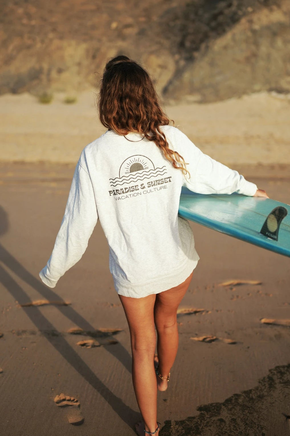 Frau mit Surfbrett in grauem Paradise & Sunset Sweater am Ozean