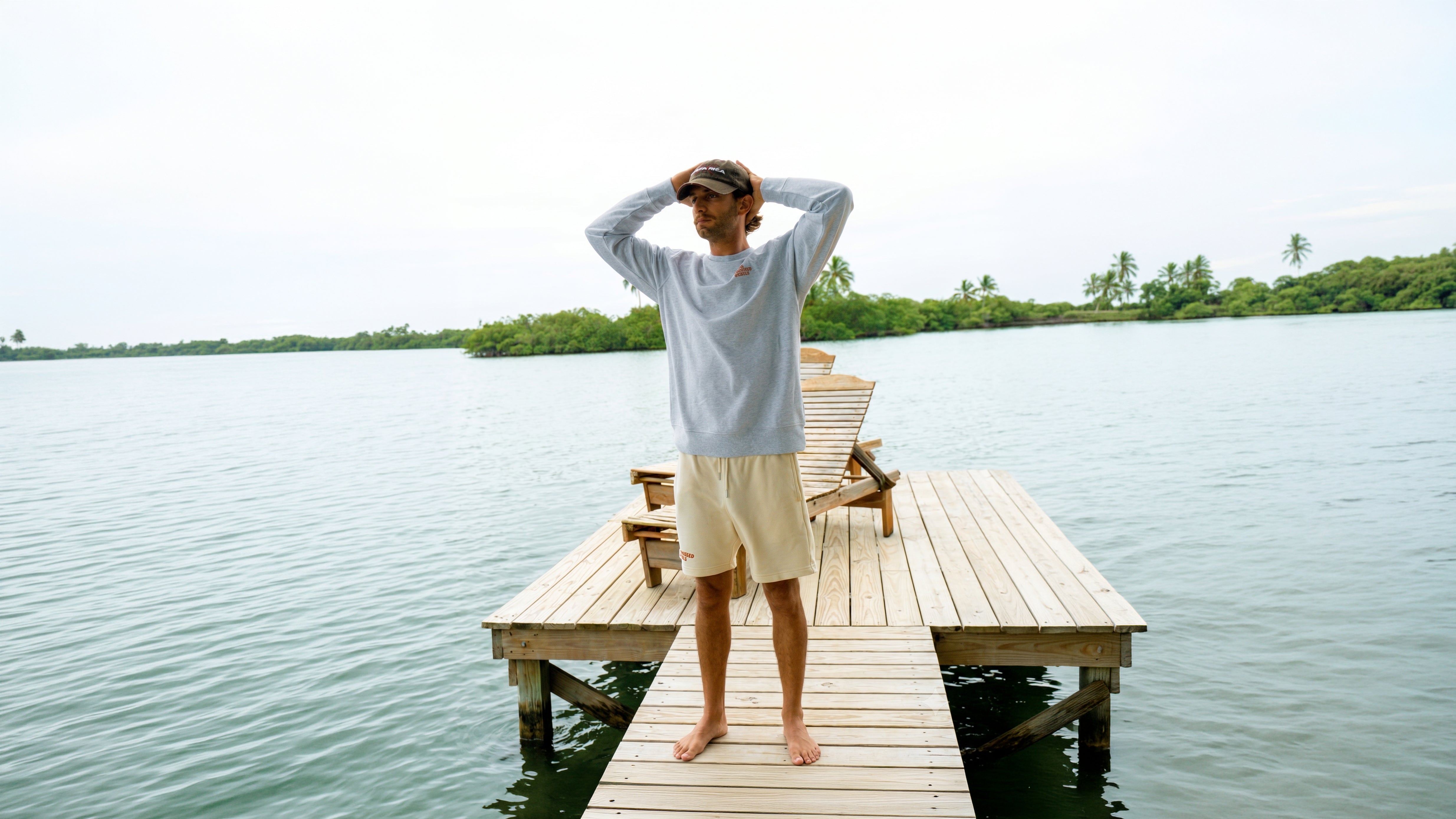 Mann trägt den Paradise & Sunset Sweater in Cool Grey auf einem Holzsteg über tropischem Wasser.