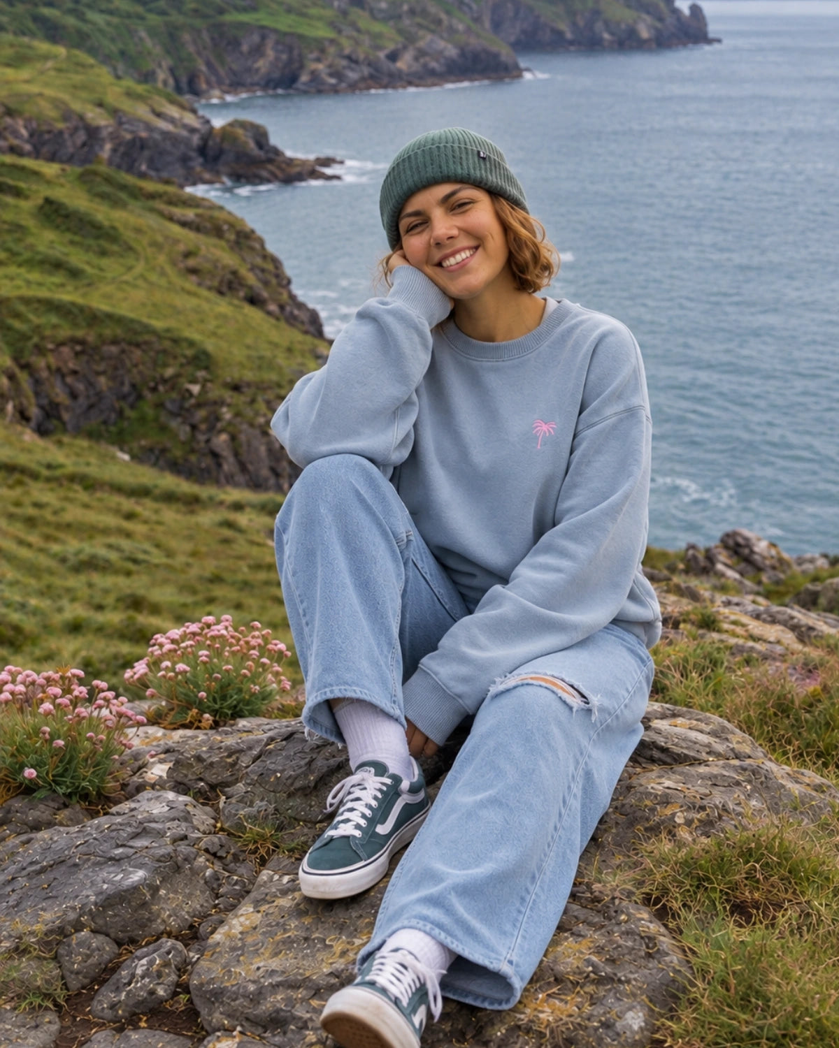 Frau sitzt im Pink Palm Oversized Vintage Sweater in Serene Blue mit pinkem Palmenstick an der Küste