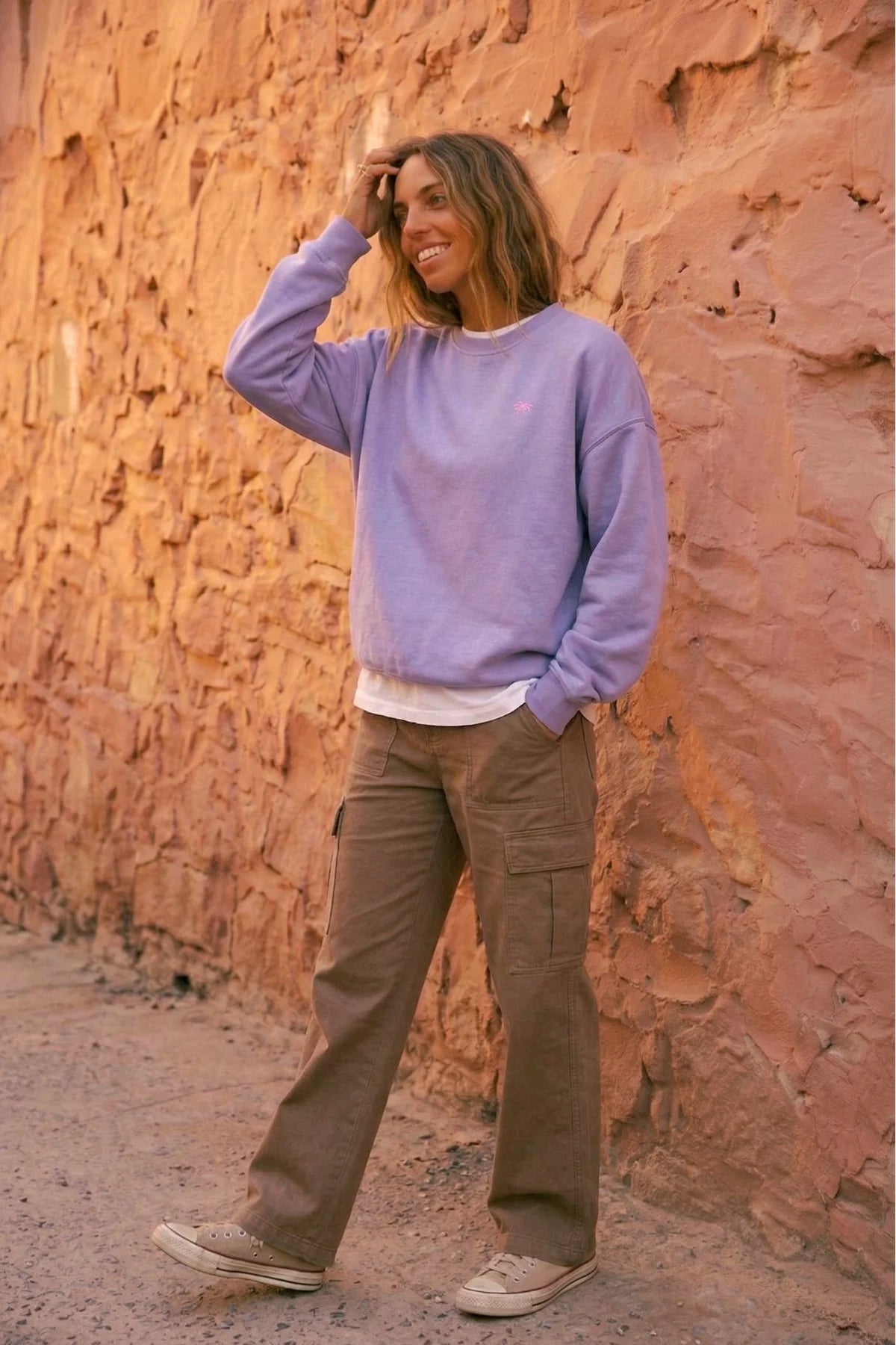 Frau lächelt und posiert im fliederfarbenen Pink Palm Oversized Vintage Sweater von The Sunkissed Rebels. Zeigt den lockeren Fit und das Palmen-Logo.