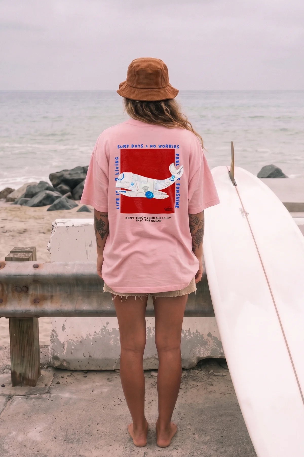 Frau mitz Surfbrett trägt pinkes Oversized T-Shirt von The Sunkissed Rebels Rückanbsicht 