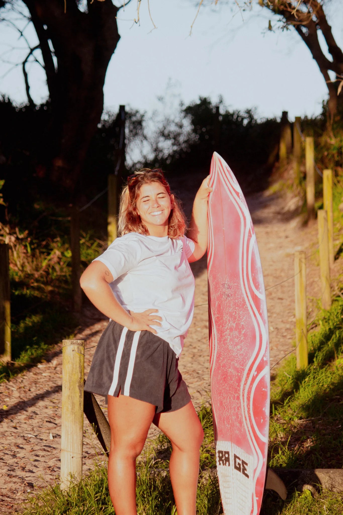 Frau mit Surfboard am Strandweg trägt weißes Rebel Elements Light T-Shirt im Sonnenuntergang
