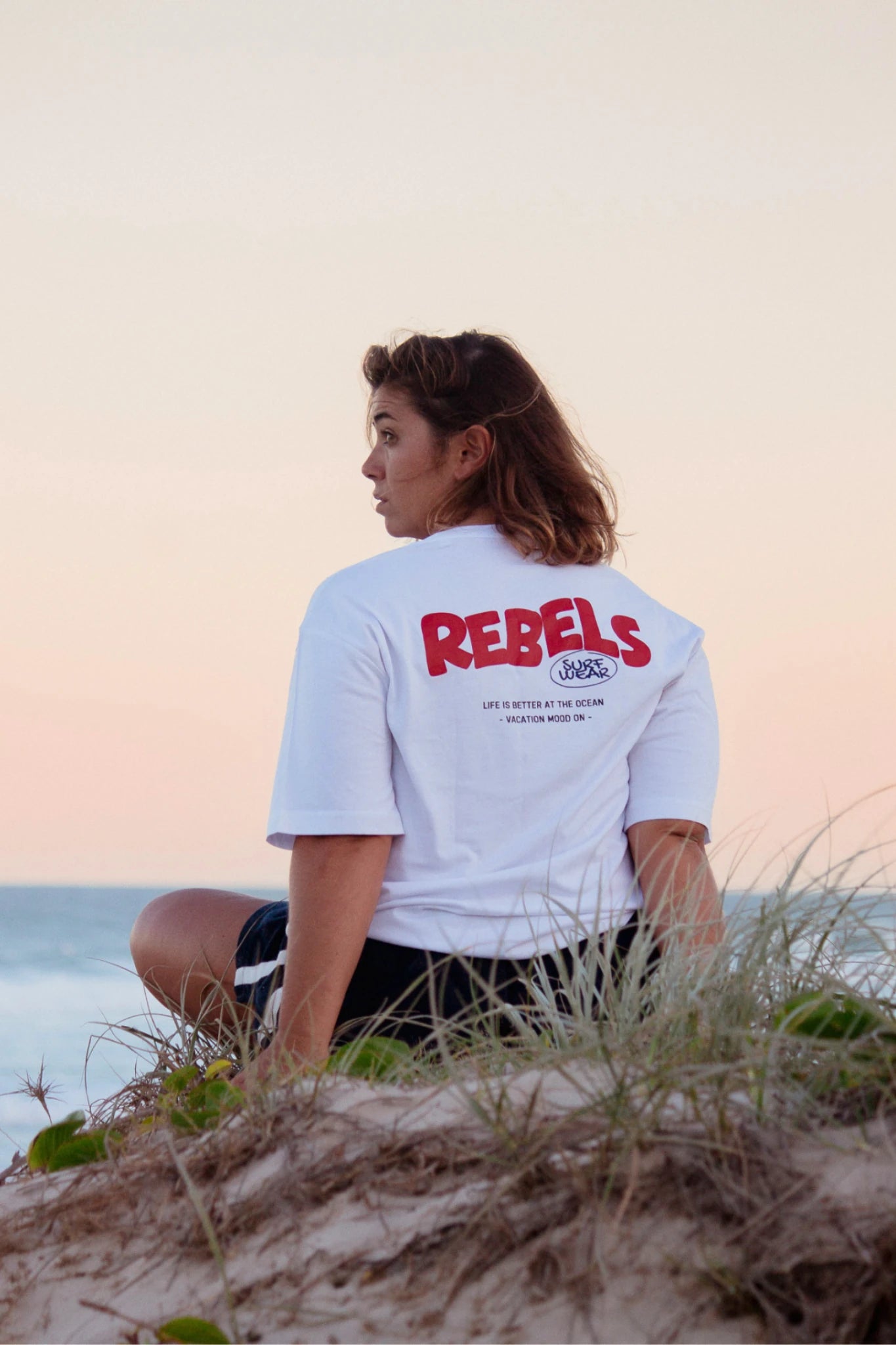 Rückansicht vom weißen Rebels Surf Wear Oversized T-Shirt mit Backprint am Meer bei Sonnenuntergang