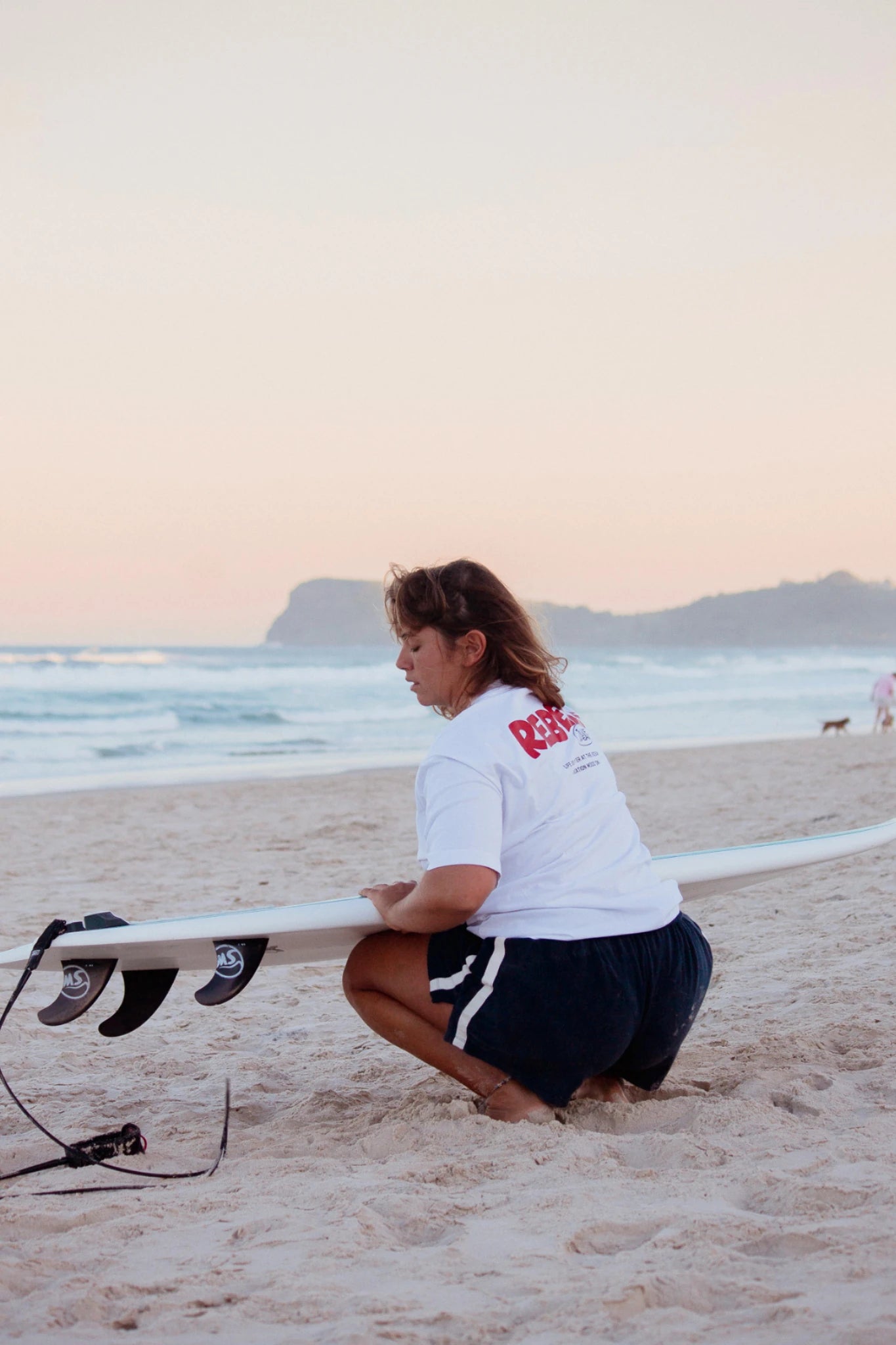 Frau am Strand mit Surfboard trägt weißes Rebels Surf Wear Oversized T-Shirt im Sonnenuntergang