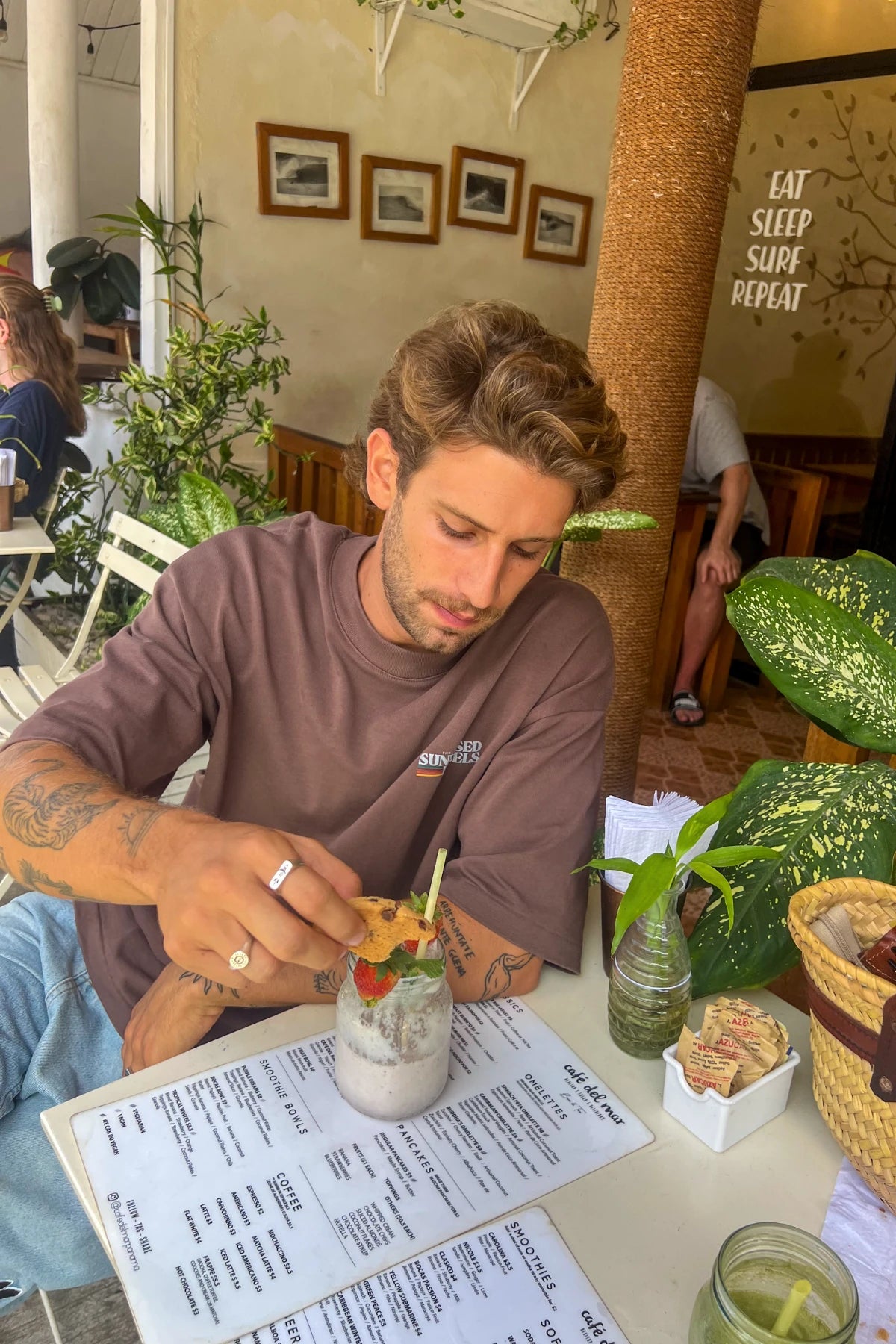 Mann trägt das Retro Vibes Heavy T-Shirt in Mocha beim Frühstück in einem tropischen Café mit Smoothie und Cookie.