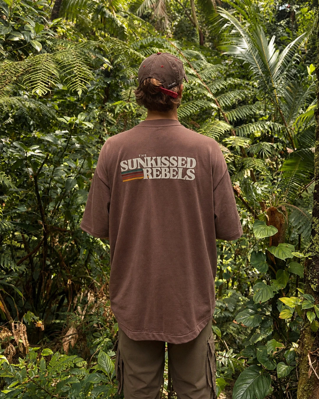 Mann trägt das Retro Vibes T-Shirt in Mocha von The Sunkissed Rebels mit großem Backprint in tropischer Umgebung.