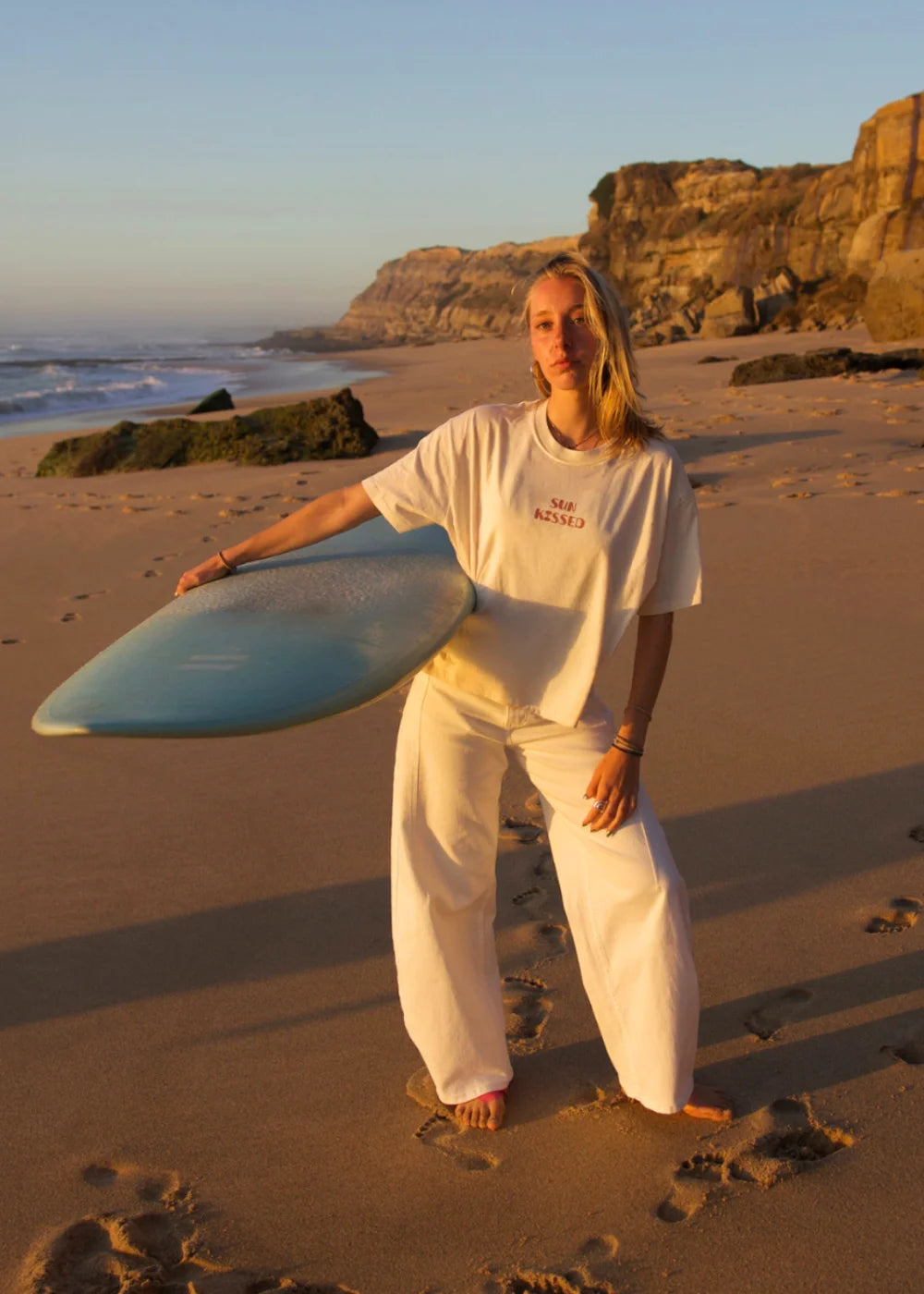 Frau trägt das weiße Sun Kissed Oversized T-Shirt von The Sunkissed Rebels mit Surfboard am Strand bei Sonnenuntergang.