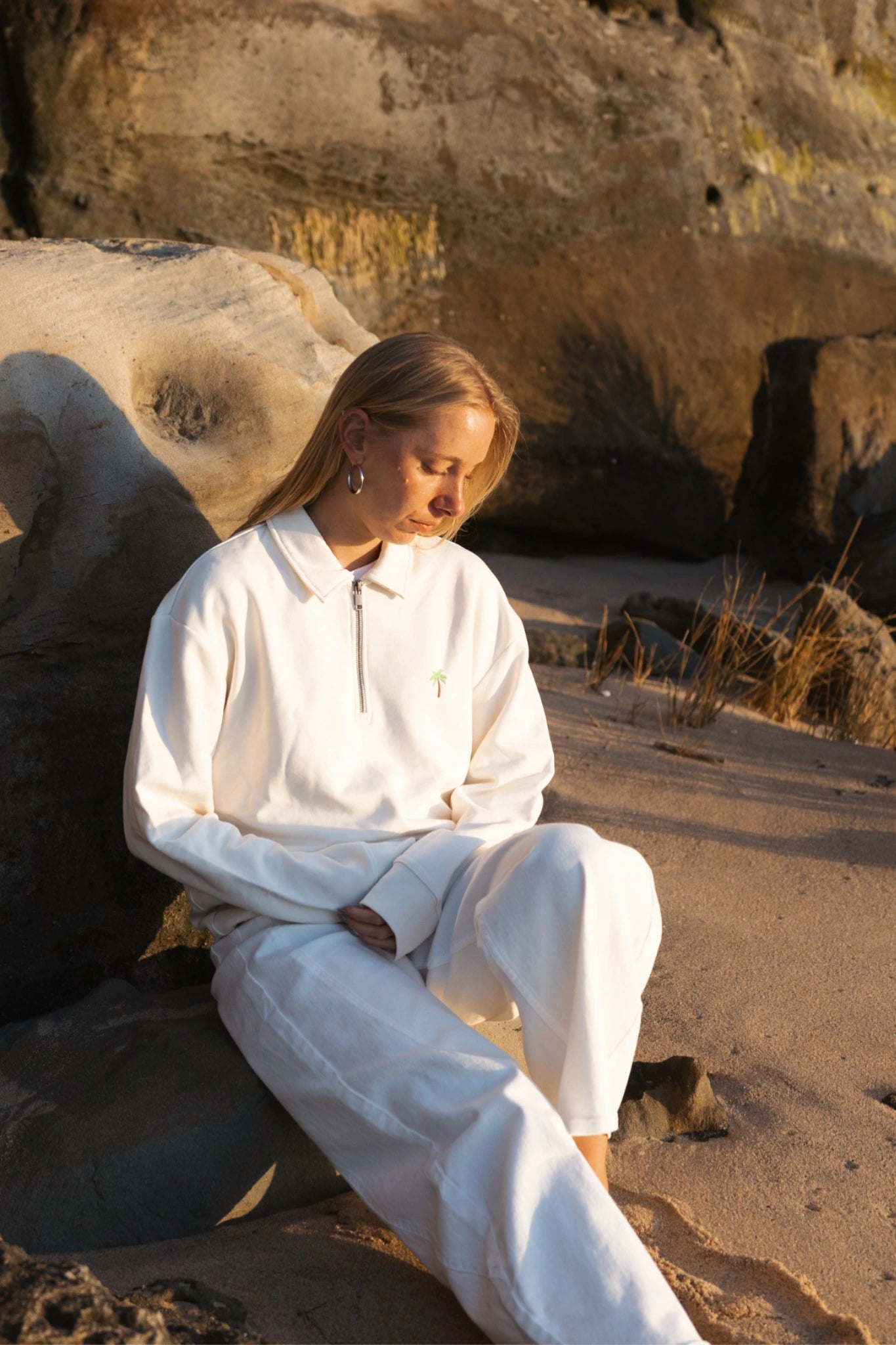 Frau sitzt am Strand an Felsen und trägt cremefarbenen Half-Zip Pullover im warmen Abendlicht