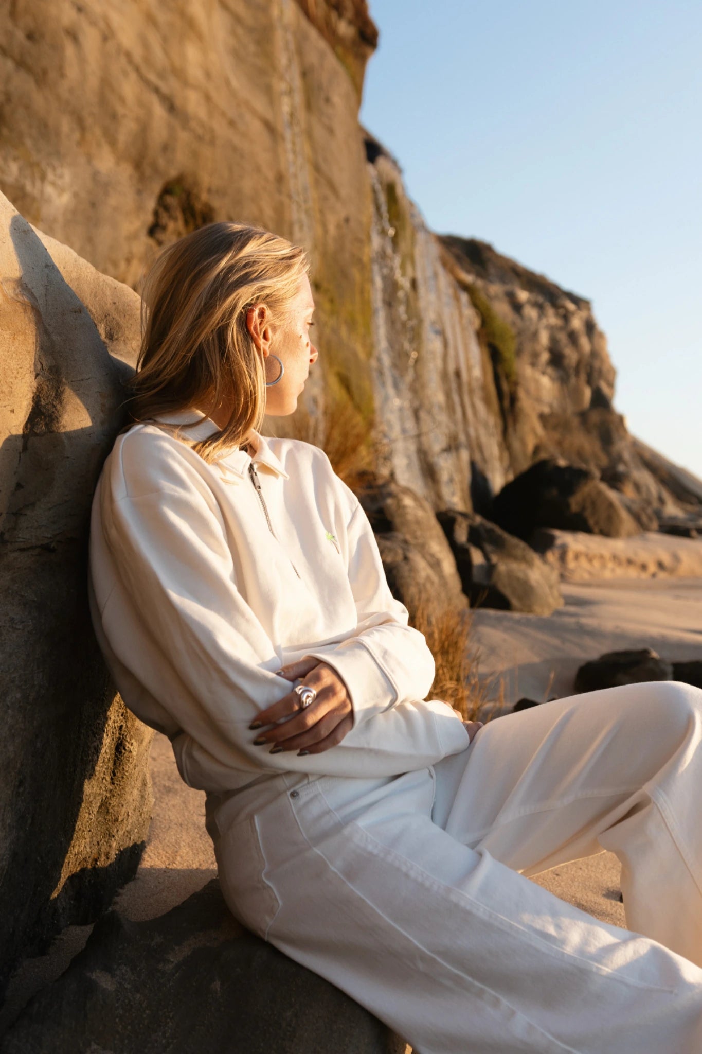 Frau lehnt an Felsen und trägt cremefarbenen Half-Zip Pullover im Sonnenuntergang am Strand
