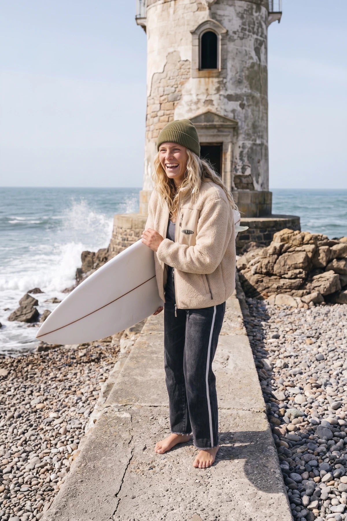Blonde Surferin mit beiger Teddy Fleece Jacke von Sunkissed Rebels und Surfboard vor einem Leuchtturm am Meer.