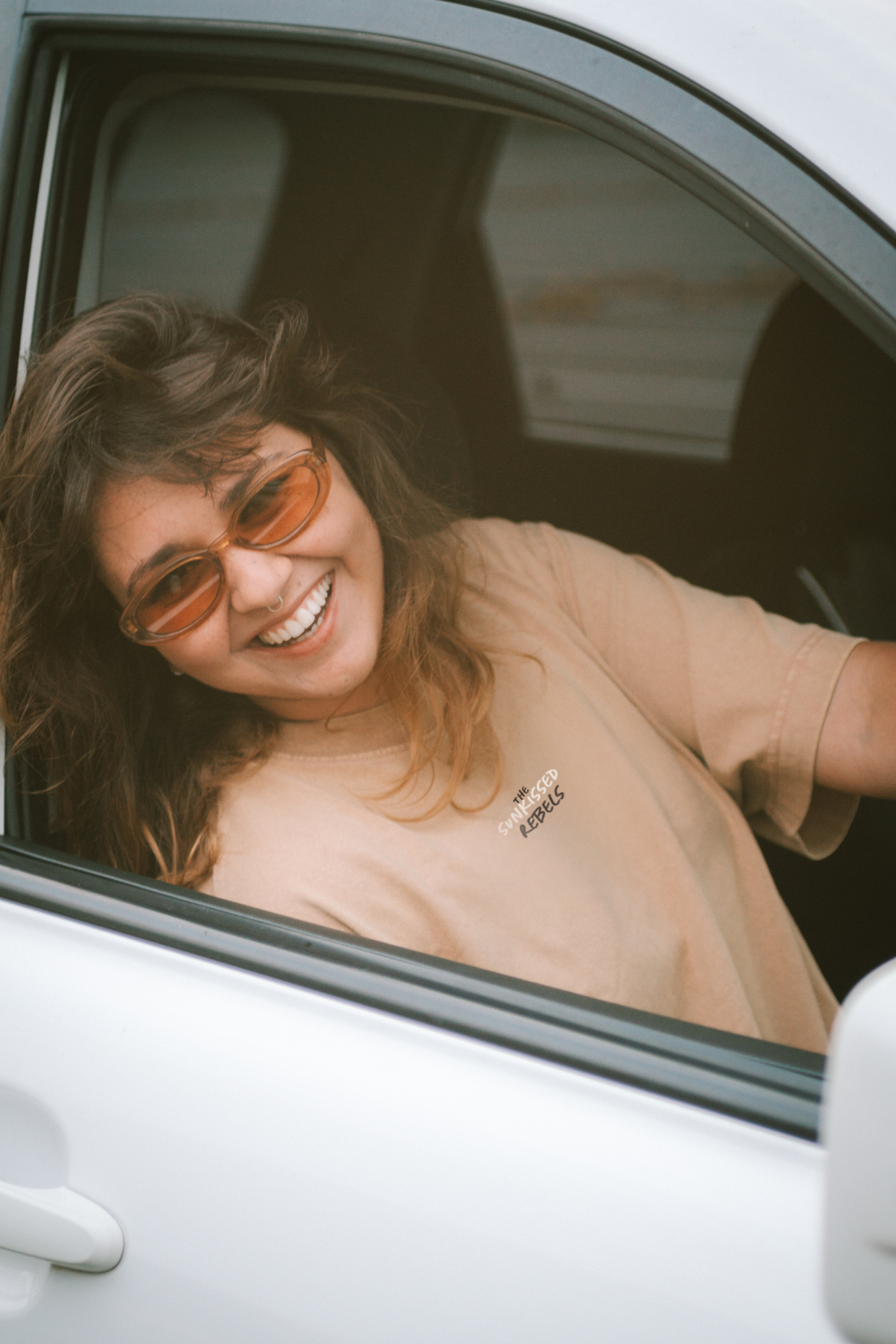 Frau sitzt im Auto, lacht in Kamera und trägt dabei ein T-Shirt von The Sunkissed Rebels. 