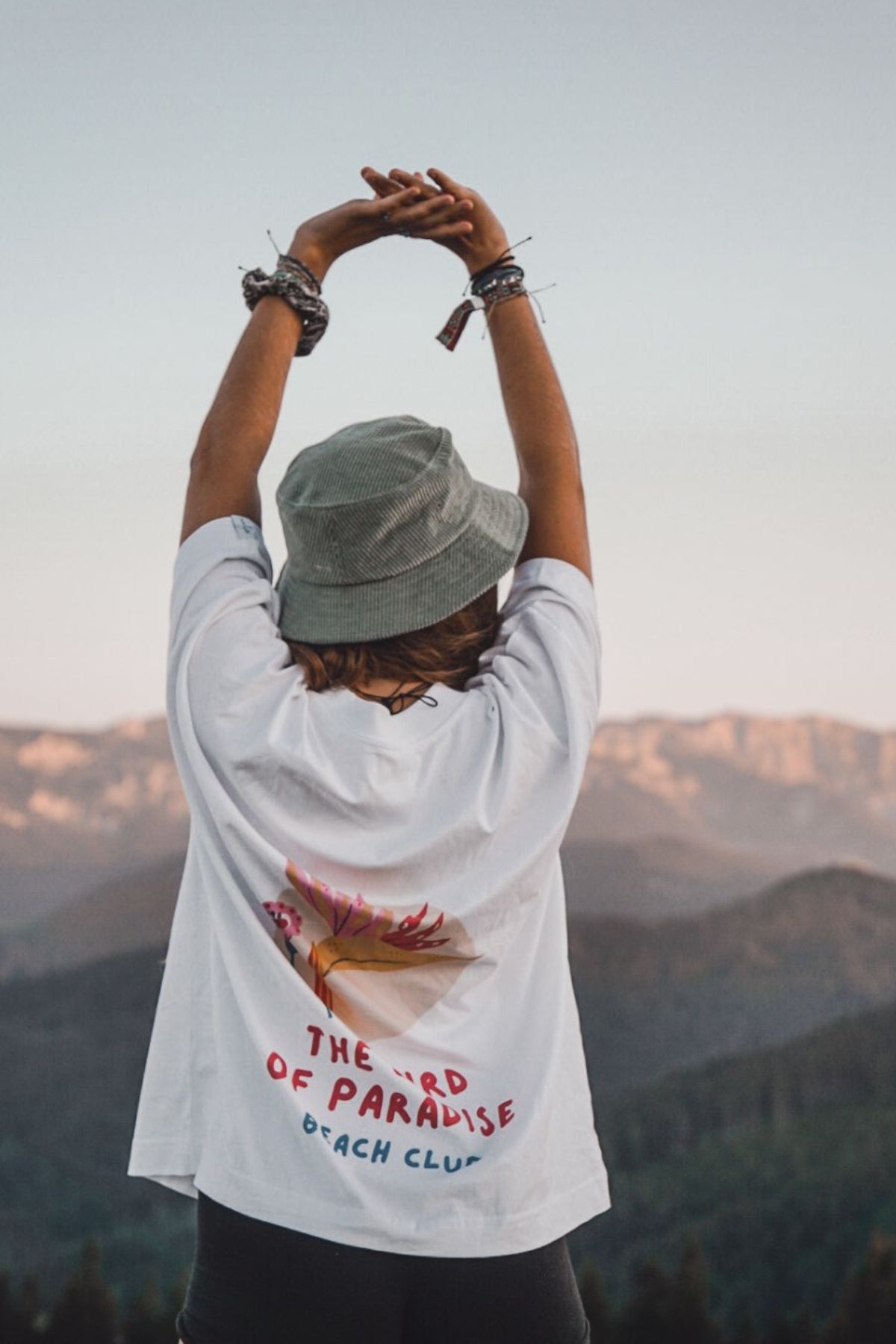 Frau steht in den Bergen, streckt die Arme aus und trägt das oversized T-Shirt Bird of Paradise in weiß von The Sunkissed Rebels. 