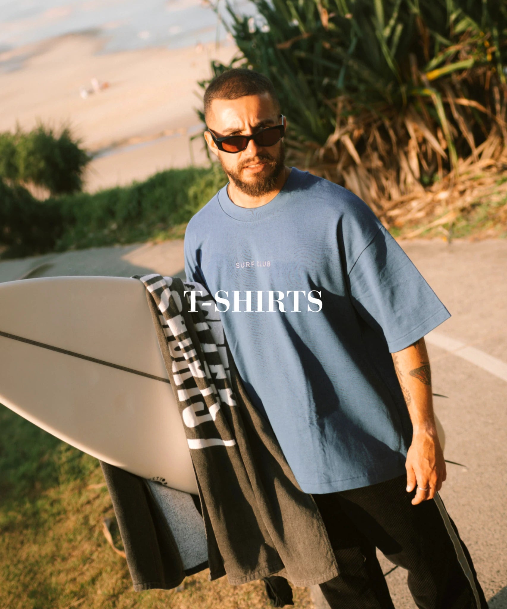 Mann mit Surfbrett trägt blaues T-Shirt im lässigen Surf Style am Strand bei Sonnenuntergang