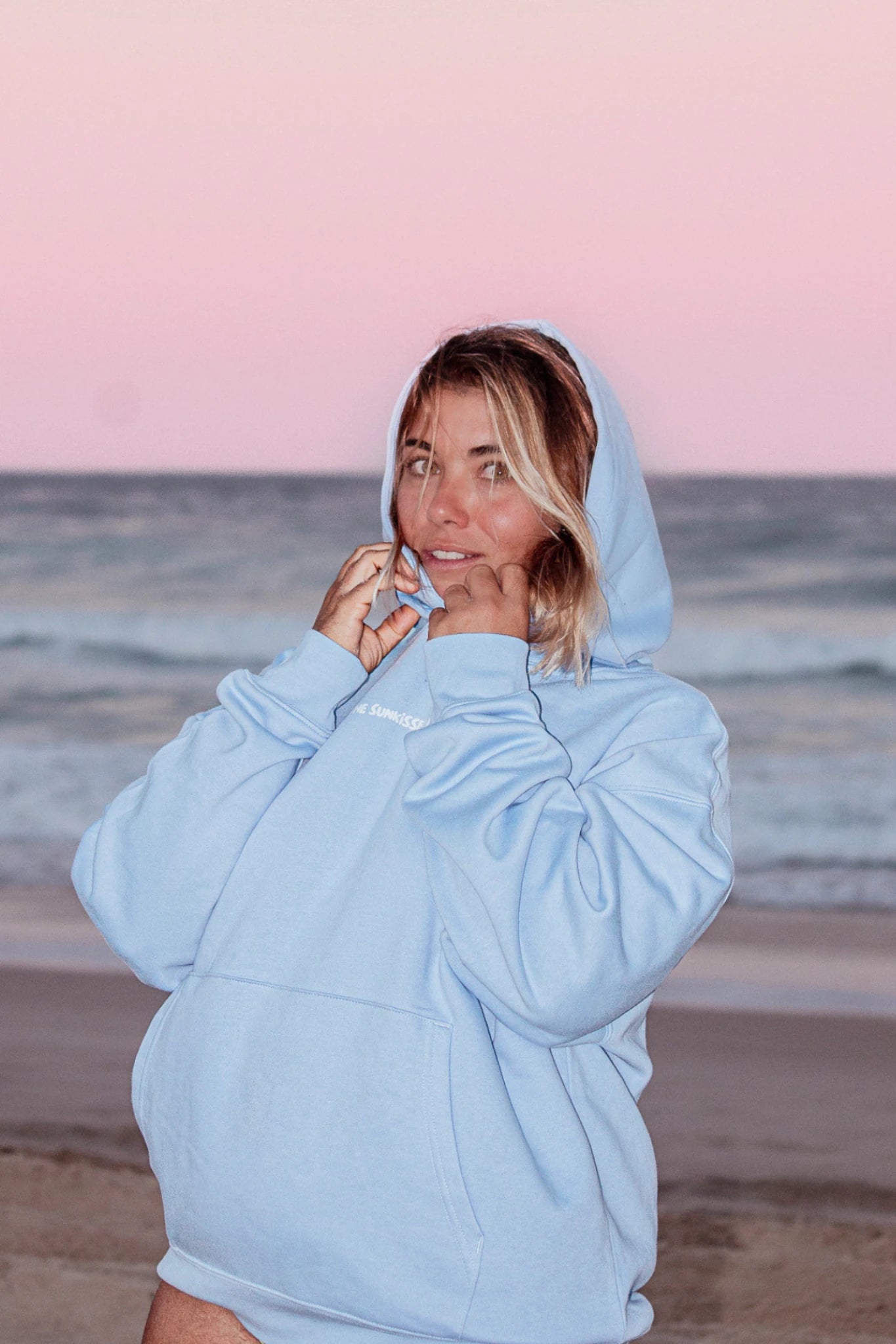 Frau lächelt im blauen Hoodie am Strand mit Ozean im Hintergrund