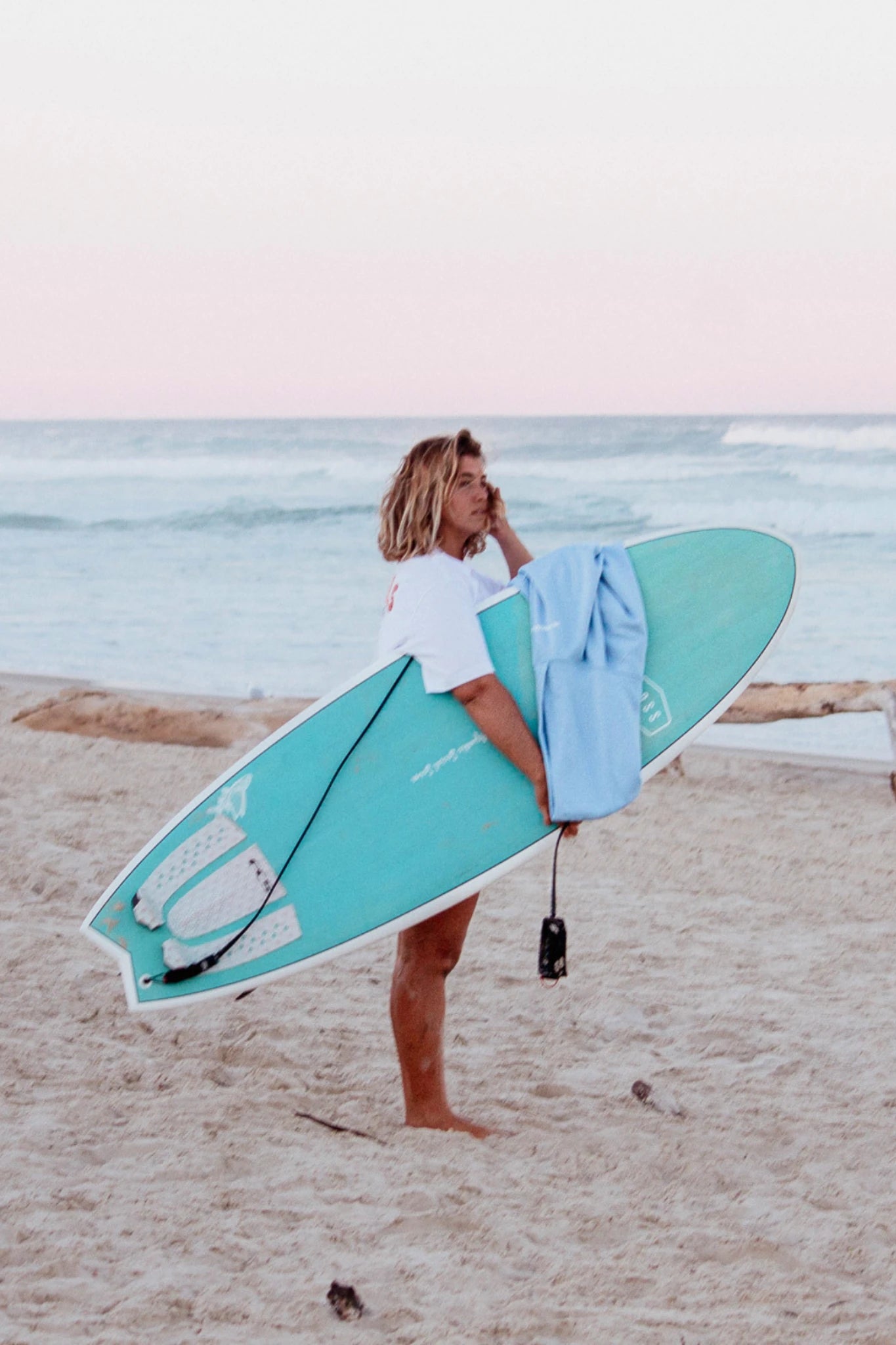 Frau im blauen Hoodie mit Surfbrett am Strand bei Sonnenuntergang