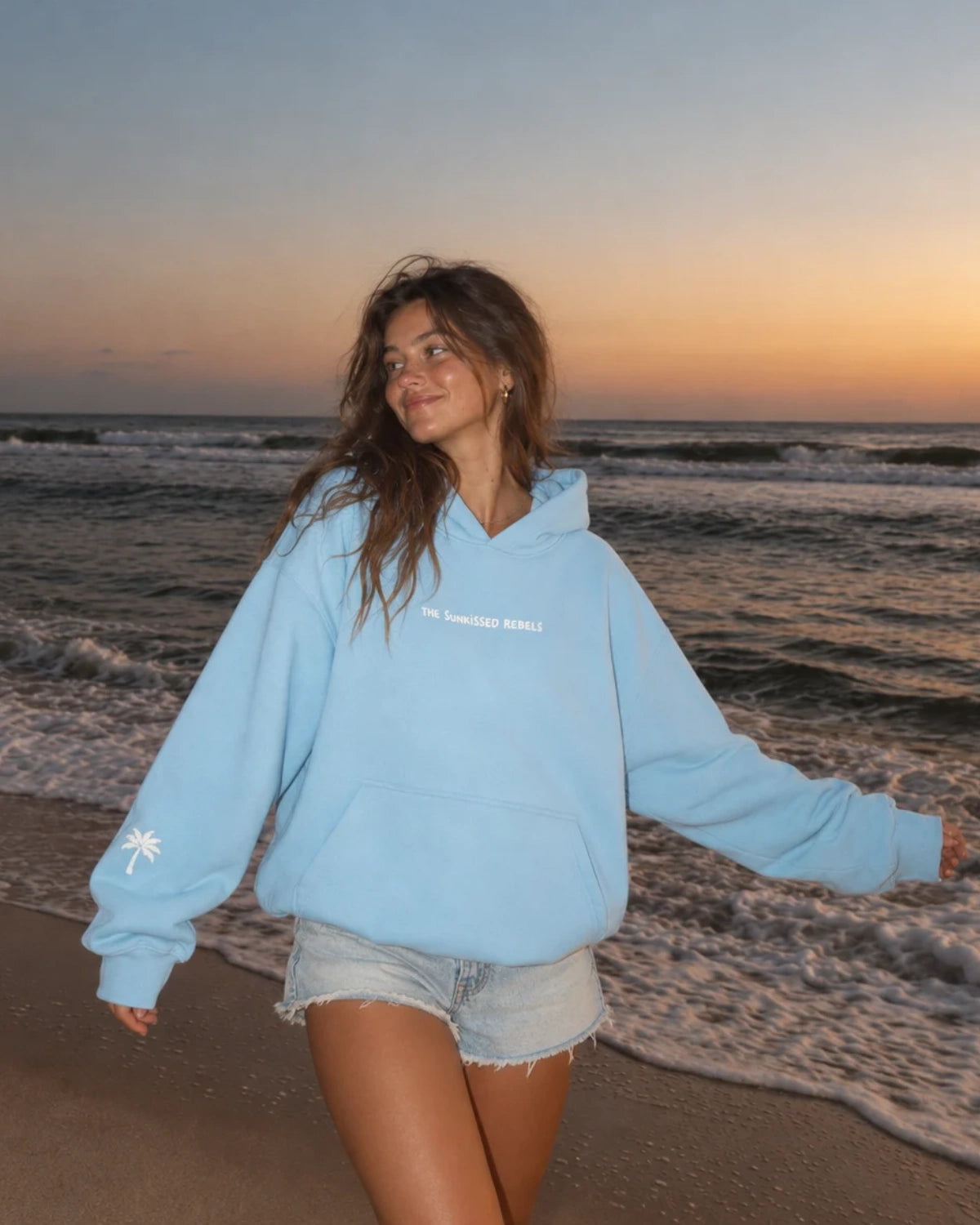 Frau trägt den hellblauen The Horizon Hoodie – Blue Soul von The Sunkissed Rebels am Strand bei Sonnenuntergang.