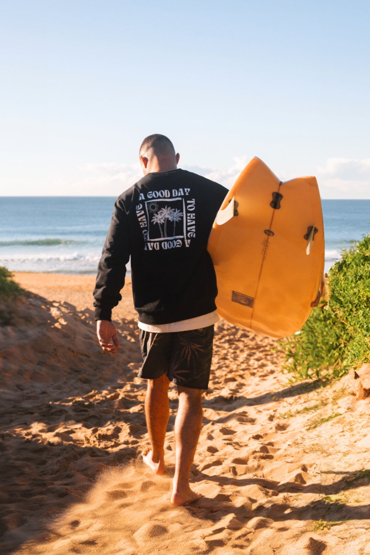Surfer trägt schwarzen Sweater mit einem Palmen Print und der Aufschrift A Good Day To Have A Good Day. 