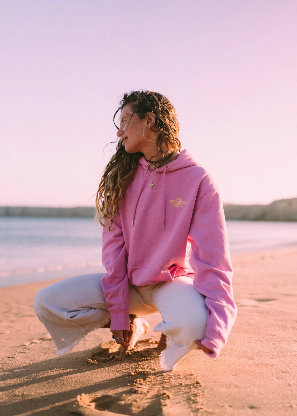 Frau trägt den pinken Oversized Hoodie von The Sunkissed Rebels an einem Sandstrand während des Sonnenuntergangs im Beach-Lifestyle.