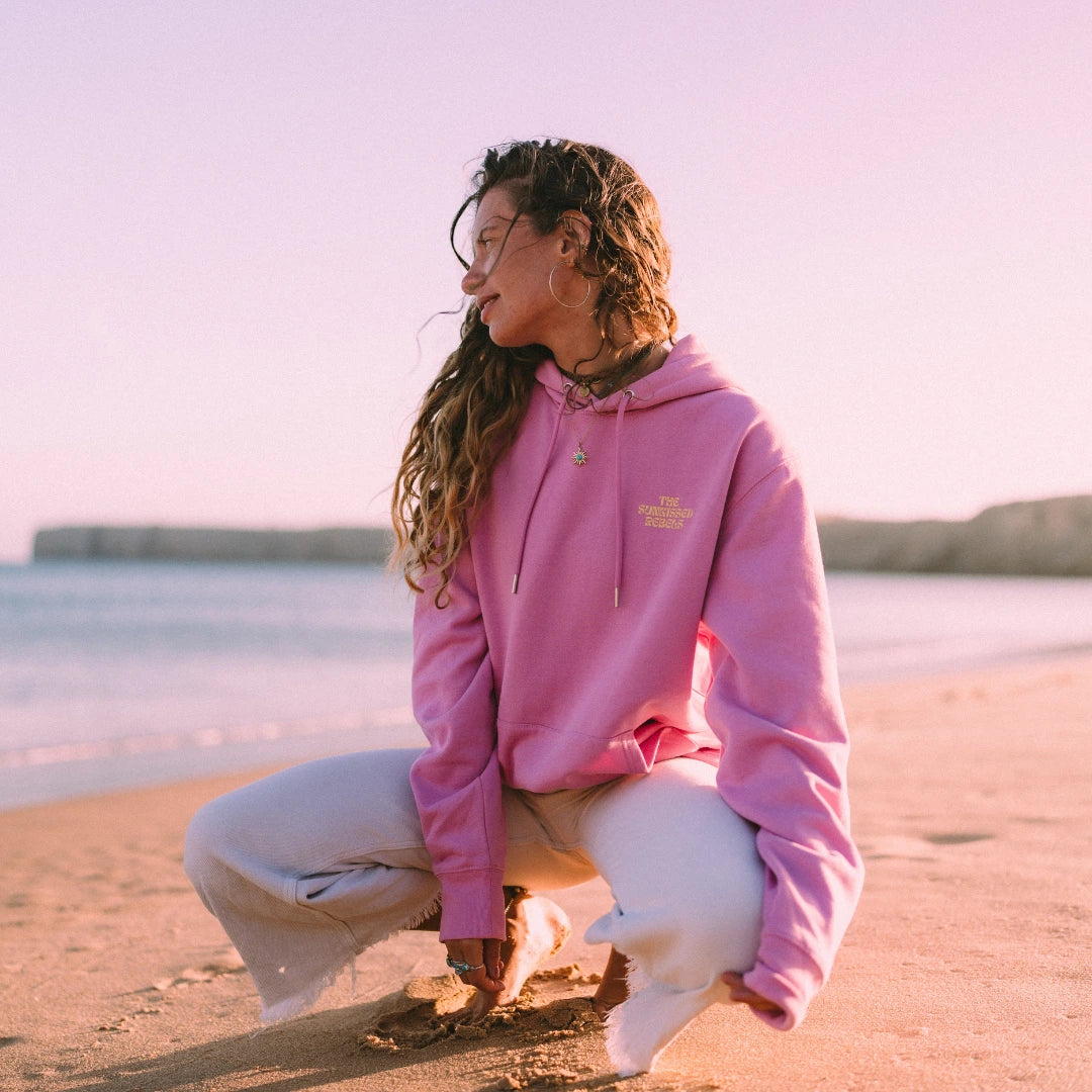 Frau trägt einen pinken The Sunkissed Rebels Hoodie am Strand bei warmem Abendlicht.