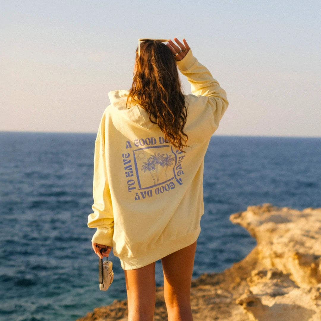 Rückansicht einer Frau im gelben The Sunkissed Rebels Hoodie mit Blick auf das Meer und die Felsküste bei Sonnenuntergang.