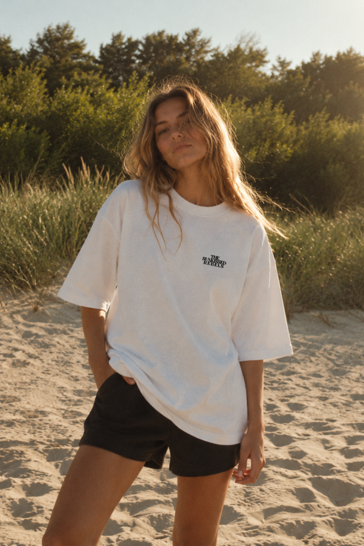 Frau am Strand im weißen Vacation Mood On Boxy Oversized T-Shirt von The Sunkissed Rebels mit schwarzem Brustlogo und weiten Ärmeln.