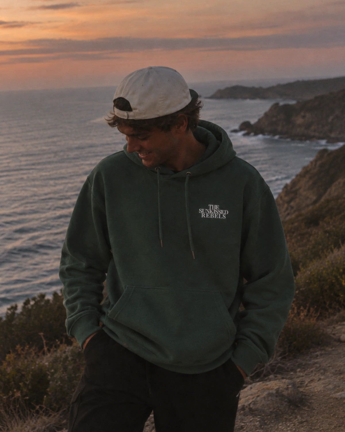 Dunkelgrüner Vacation Mood On Hoodie in Green Bay aus Bio-Baumwolle mit weißem The Sunkissed Rebels Bruststick an der Küste