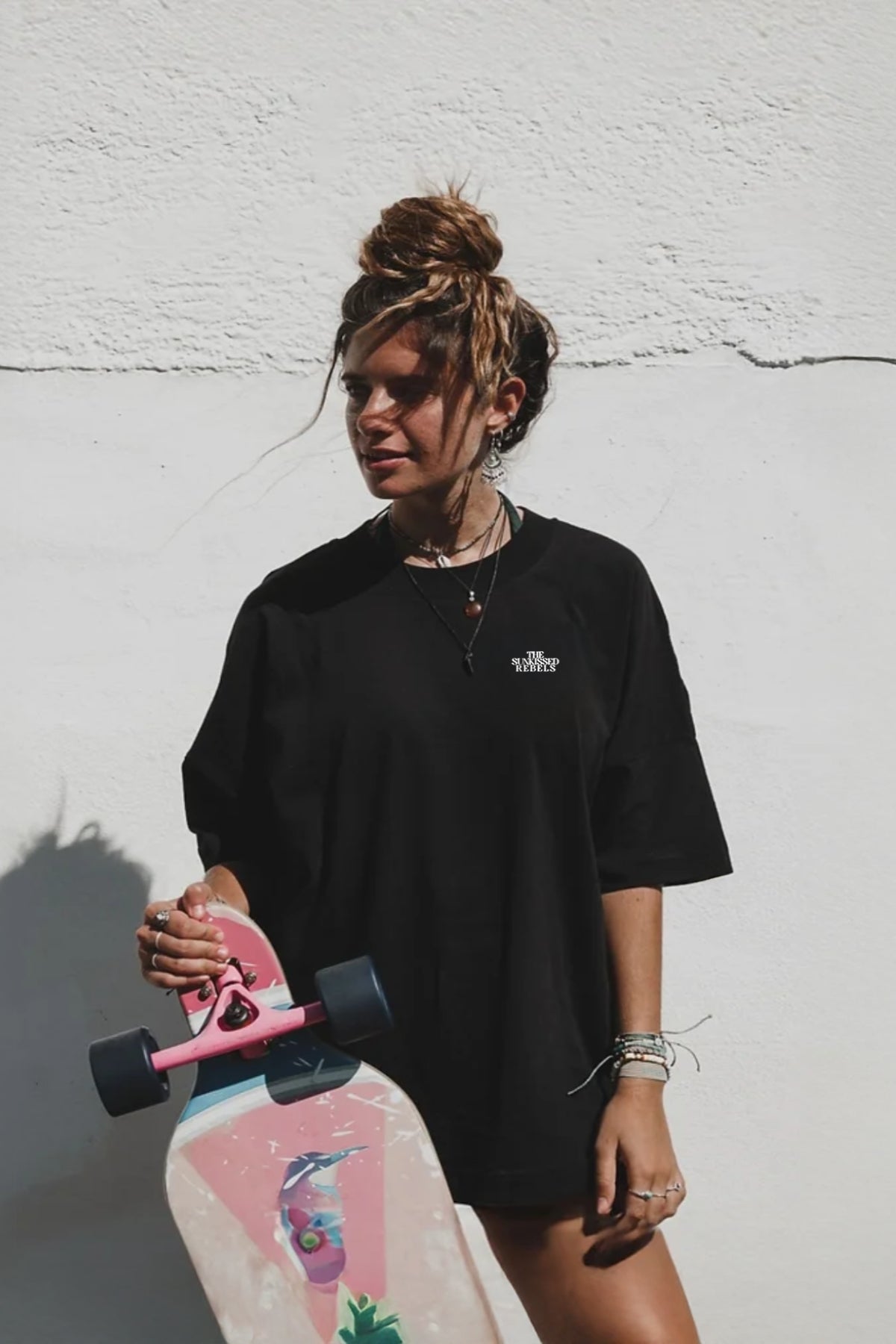 Frau trägt das Oversized Vacation Mood On T-Shirt in schwarz von The Sunkissed Rebels und hält ein Longboard in der Hand. 