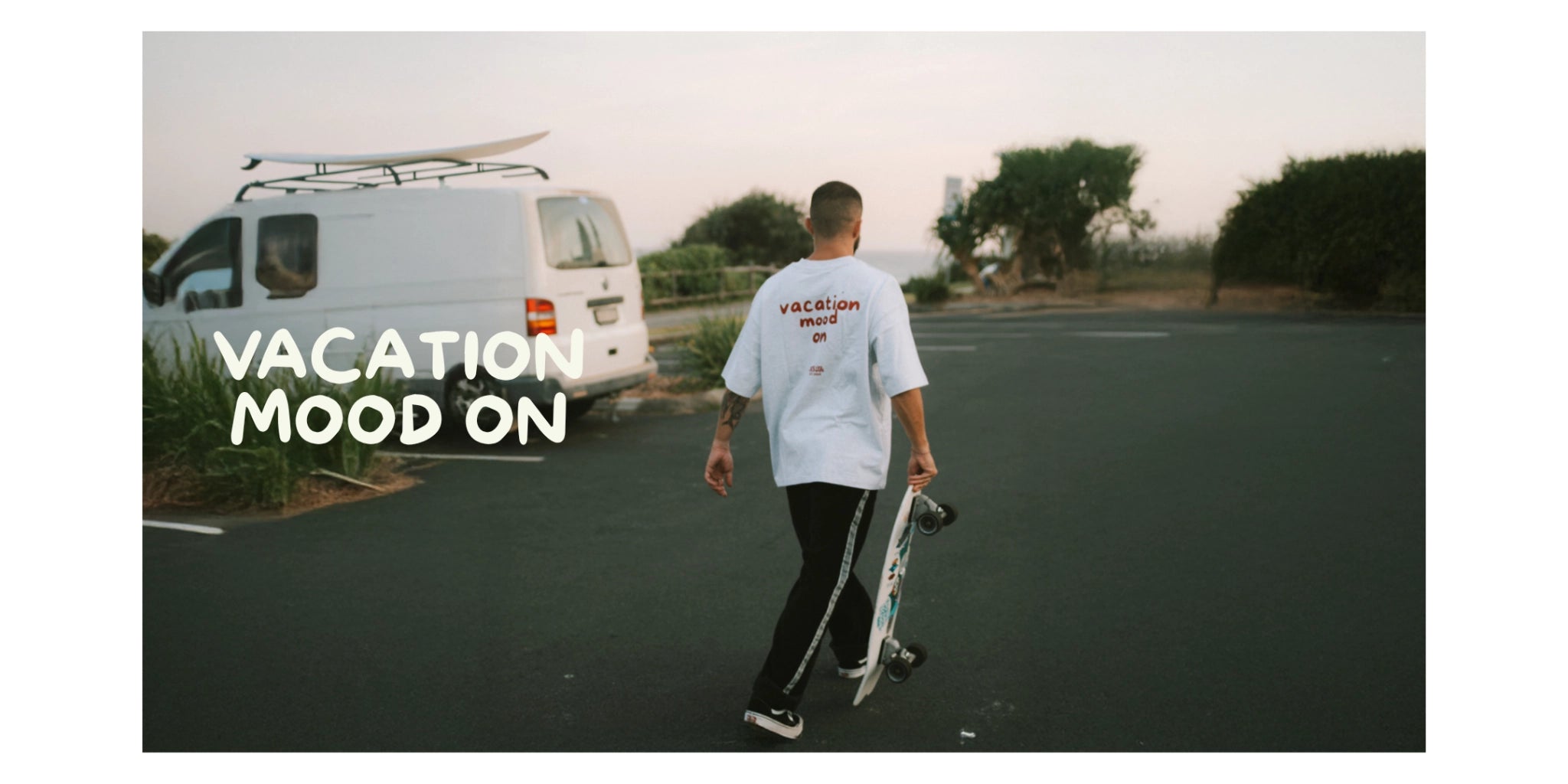 Mann mit skatebopard auf einem Parkplatz trägt graues Oversiuzed T-shirt Vacation Mood On und Jogginghose dazu Vans