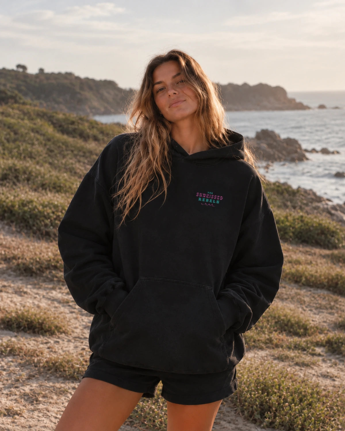 Black – Frontansicht
Frau im Vacation Vibes Oversized Hoodie in Schwarz mit farbigem The Sunkissed Rebels Brustprint an der Küste
