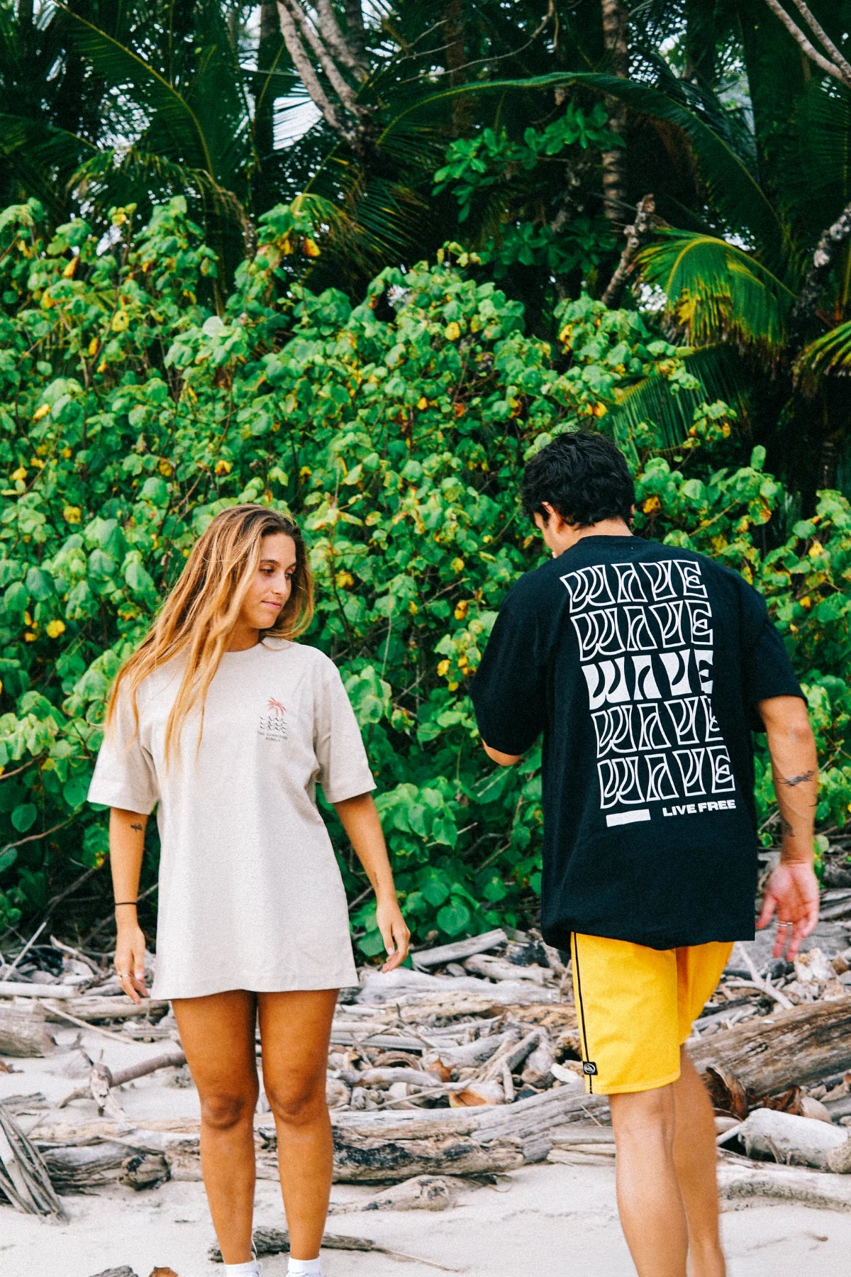 Zwei Personen tragen das Waves & Palm Trees Oversized T-Shirt in Sand und Schwarz beim Spaziergang an einem tropischen Strand mit Palmen.