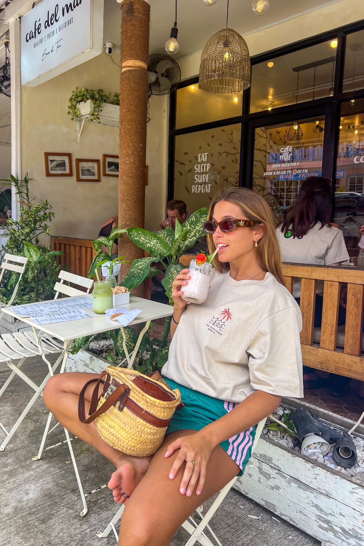 Frau trägt das Waves & Palm Trees Oversized T-Shirt in Sand beim Frühstück in einem tropischen Café mit Smoothie und Cookie.