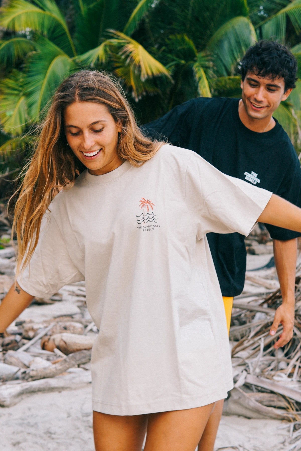 Detailaufnahme des Waves & Palm Trees Oversized T-Shirts in Sand mit Brustprint vor tropischer Strandkulisse.