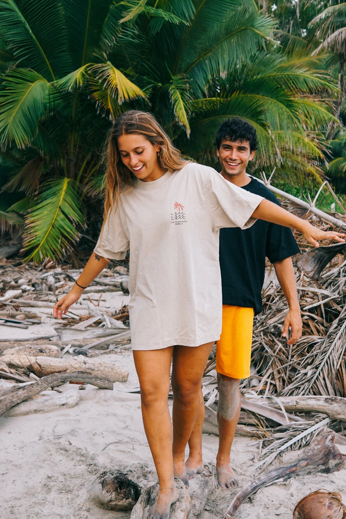 Lifestyle-Aufnahme des Waves & Palm Trees Oversized T-Shirts in Sand an einem tropischen Strand mit Palmen und Treibholz