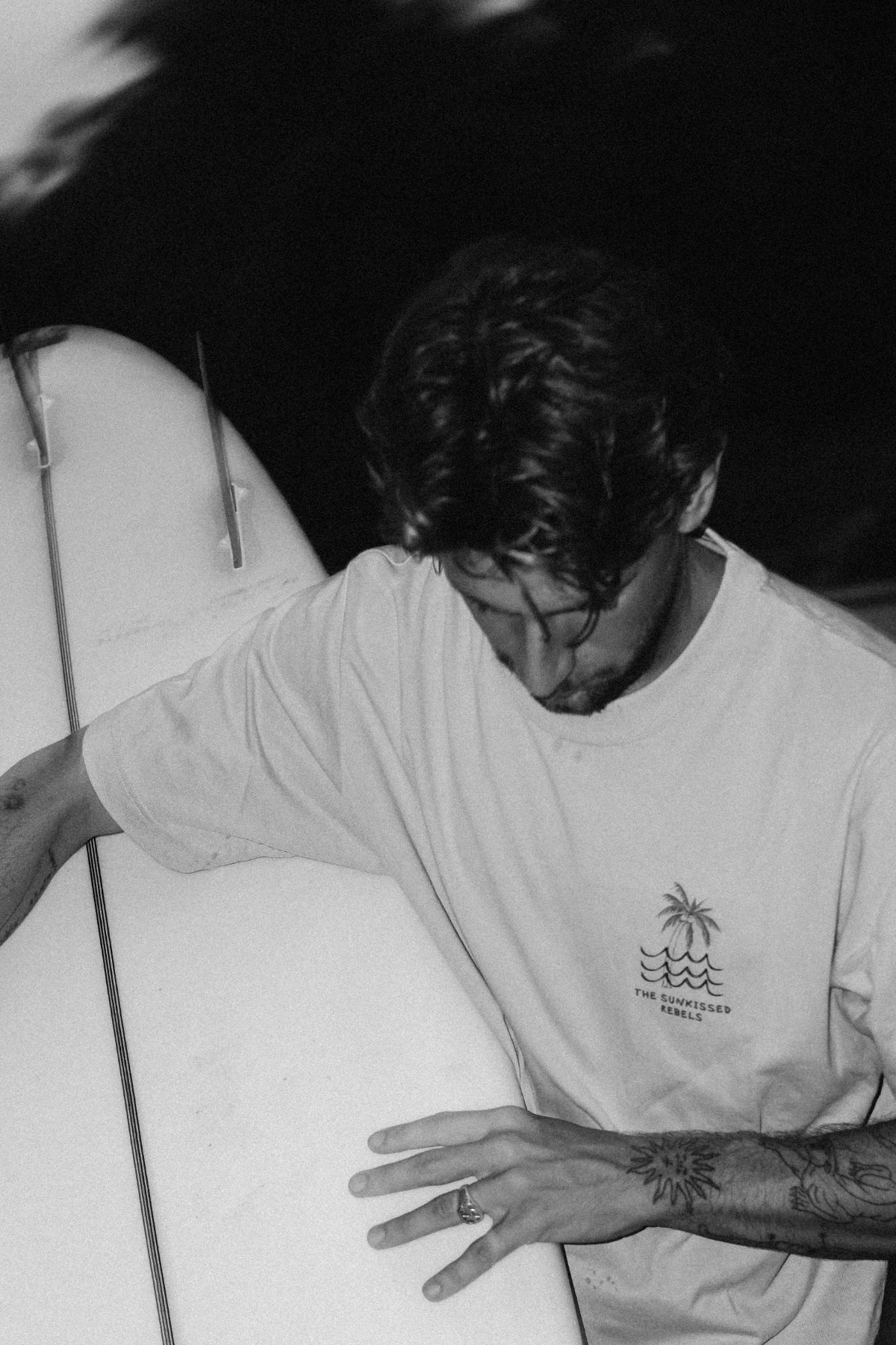 Schwarz-Weiß-Aufnahme eines Surfers mit dem Waves & Palm Trees Oversized T-Shirt in Sand und Surfboard am Strand.