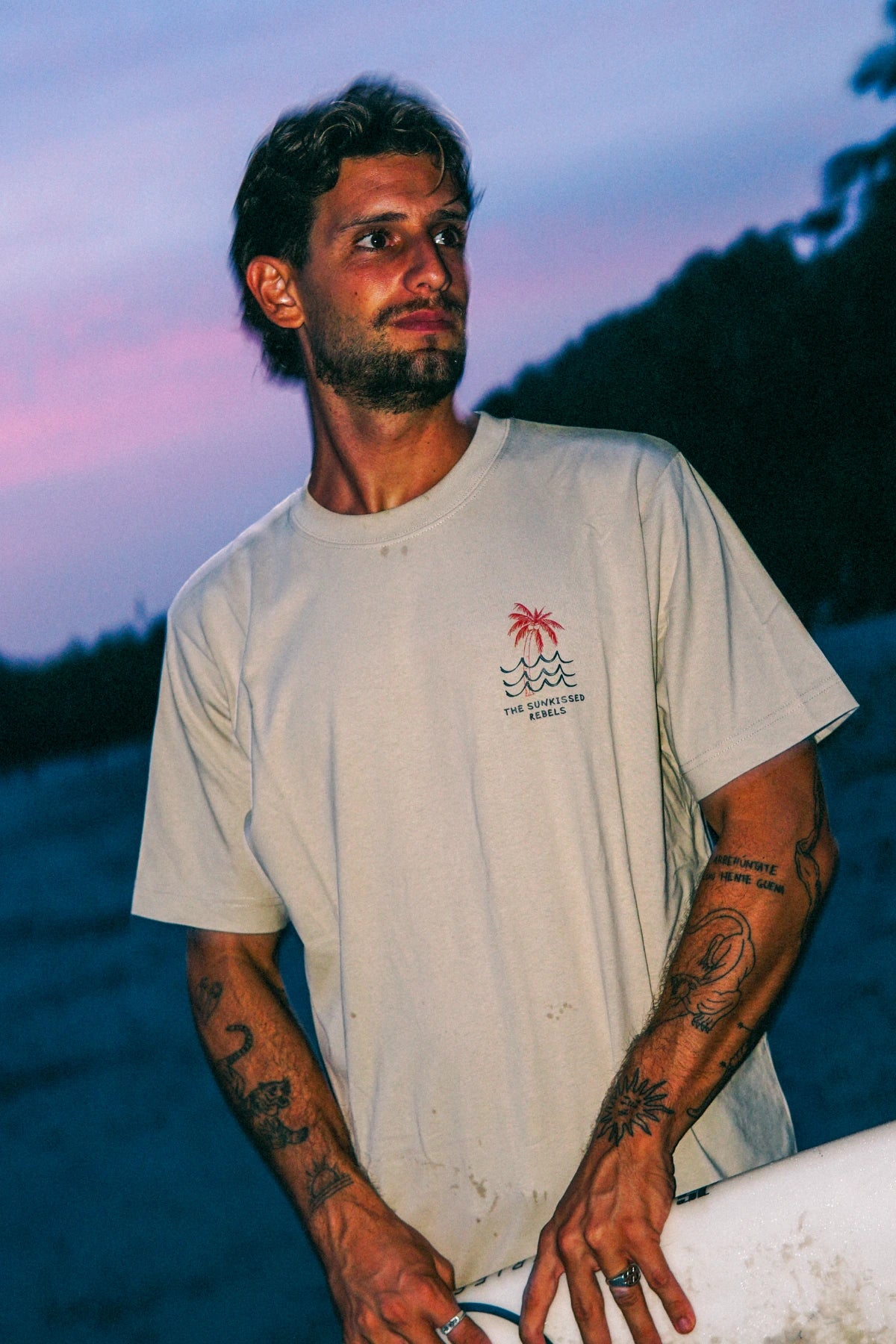 Mann trägt das Waves & Palm Trees Oversized T-Shirt in Sand mit Brustprint und Surfboard bei Sonnenuntergang am Strand.