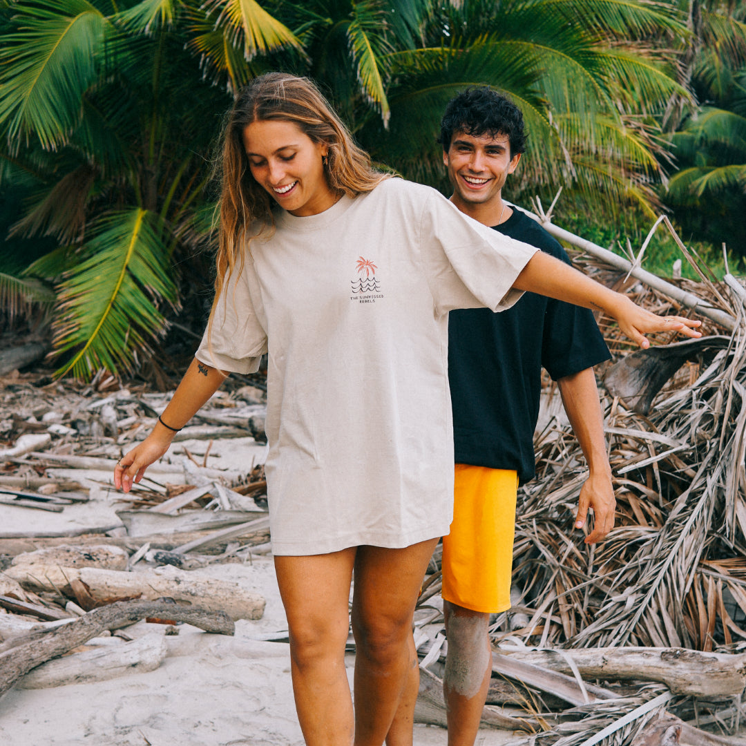 Frau trägt das Waves & Palm Trees Oversized T-Shirt in Sand von The Sunkissed Rebels an einem tropischen Strand vor Palmen.