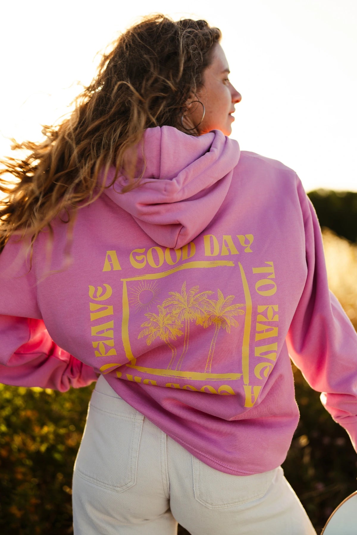 Frau trägt den A Good Day Hoodie Bubble Pink und läuft mit einem Skateboard auf einem sonnigen Küstenweg.