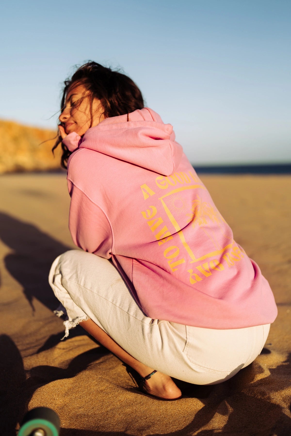 Rückenansicht des A Good Day Hoodie Bubble Pink mit großem Rückenprint am Sandstrand bei Sonnenuntergang.