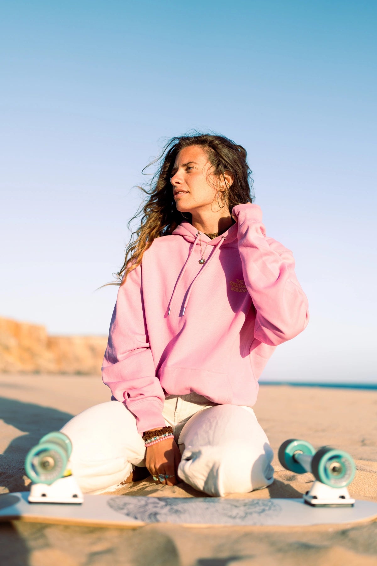 Frau trägt den A Good Day Hoodie Bubble Pink am Strand bei warmem Sonnenlicht.