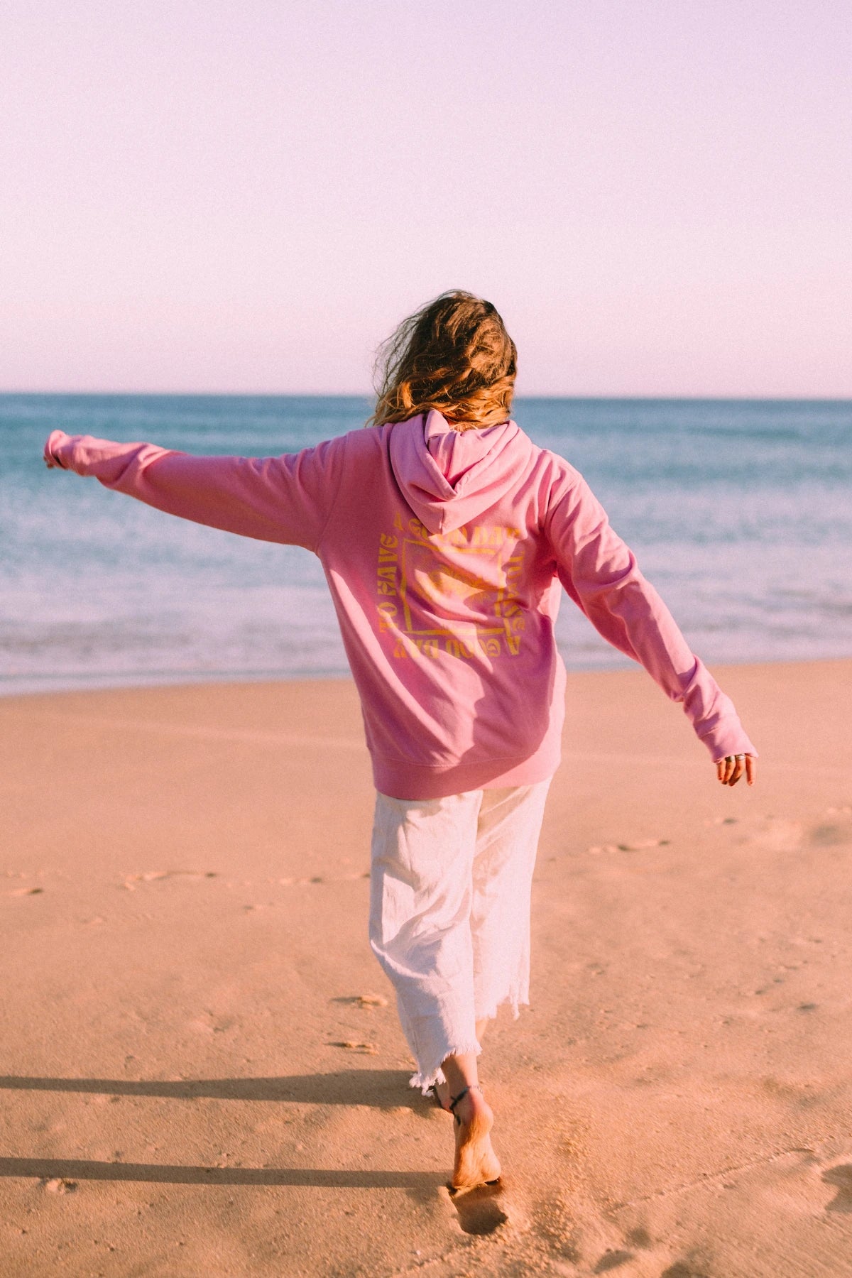 Frau trägt den A Good Day Hoodie in Bubble Pink und läuft barfuß am Strand entlang bei Sonnenuntergang.