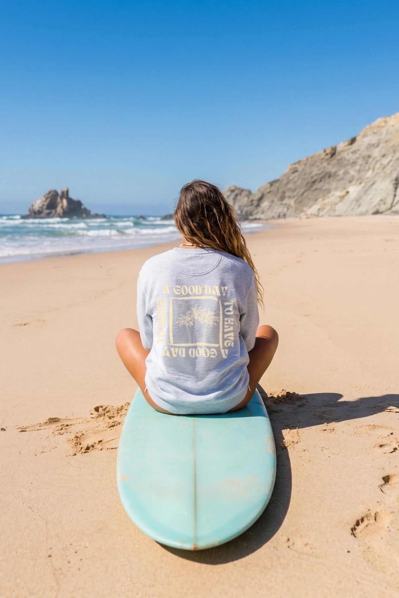Rückansicht einer Frau am Strand sitzt auf Surfboard und trägt grauen A Good Day Sweater mit Rückenprint – lässiger Beach Look